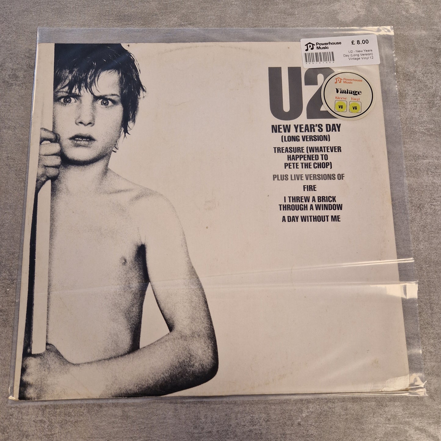 U2 - New Years Day (Long Version) Vintage Vinyl 12" Single A U2 'New Year's Day' vinyl record sleeve in a clear plastic protective case, photographed on a gray textured surface. The cover features a black-and-white portrait of a shirtless young U2 member and track listing text on the right.