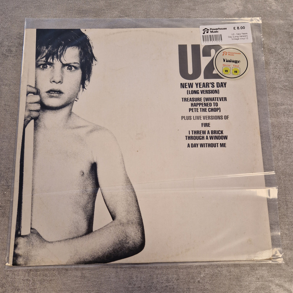 U2 - New Years Day (Long Version) Vintage Vinyl 12" Single A U2 'New Year's Day' vinyl record sleeve in a clear plastic protective case, photographed on a gray textured surface. The cover features a black-and-white portrait of a shirtless young U2 member and track listing text on the right.