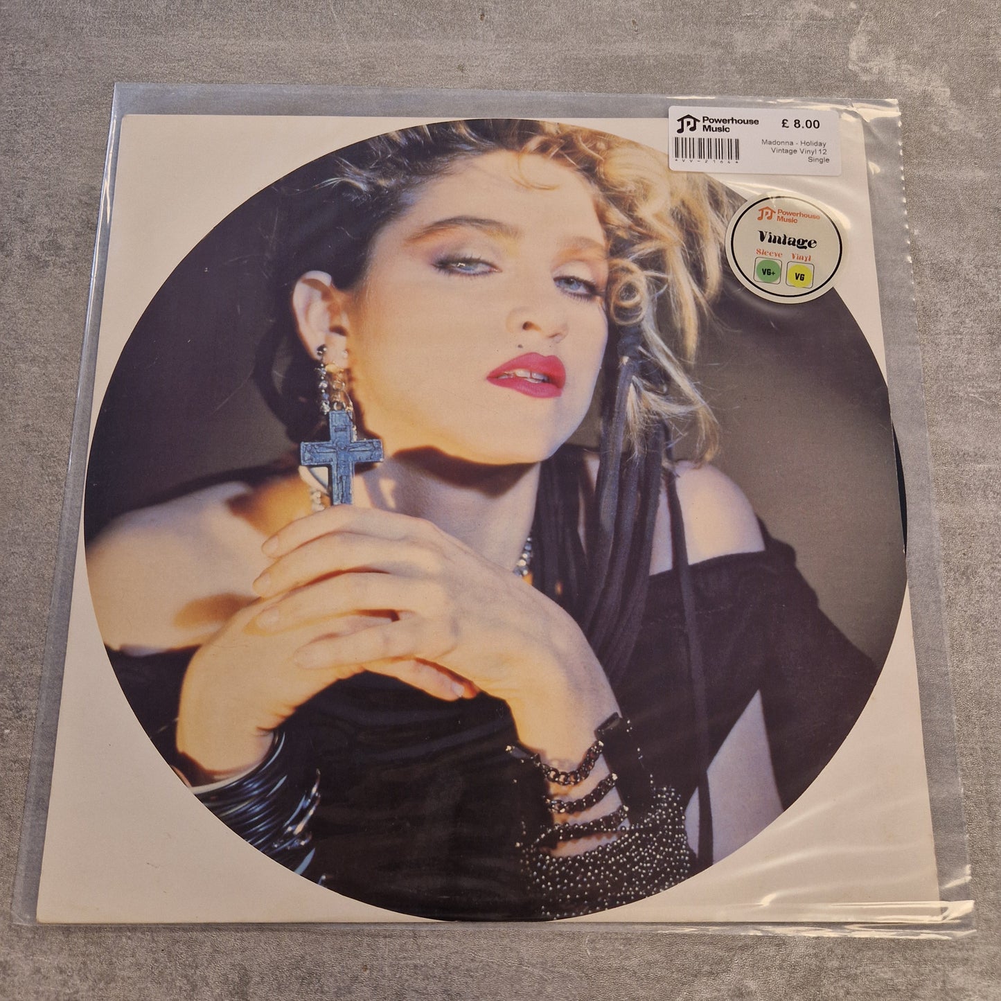 Madonna - Holiday Vintage Vinyl 12" Single A square vinyl record sleeve in plastic shrink-wrap, featuring a circular portrait of Madonna holding a cross. The sleeve is photographed on a gray textured surface, with a barcode sticker and a Vintage Records label visible on the top right.