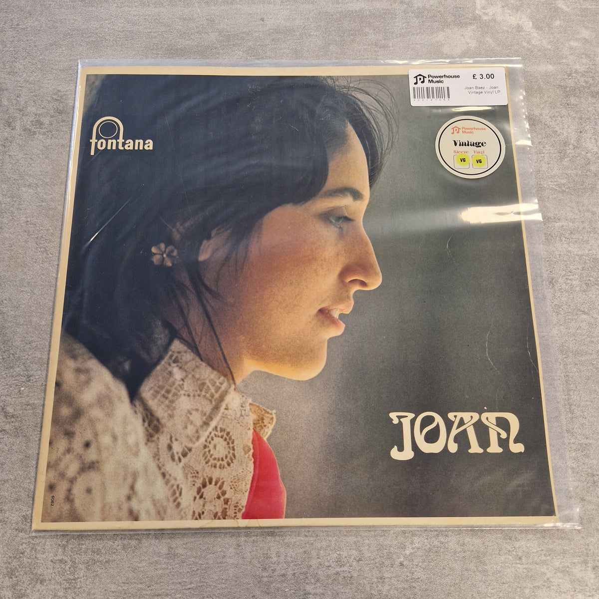Joan Baez - Joan Vintage Vinyl LP A vinyl record sleeve for Joan's album 'Fontana' is shown in its clear plastic shrink-wrap, lying on a gray textured surface. The cover features a profile portrait of a woman with dark hair, a cream lace pattern, and a red shirt, with the text 'fontana' and 'JOAN' visible.