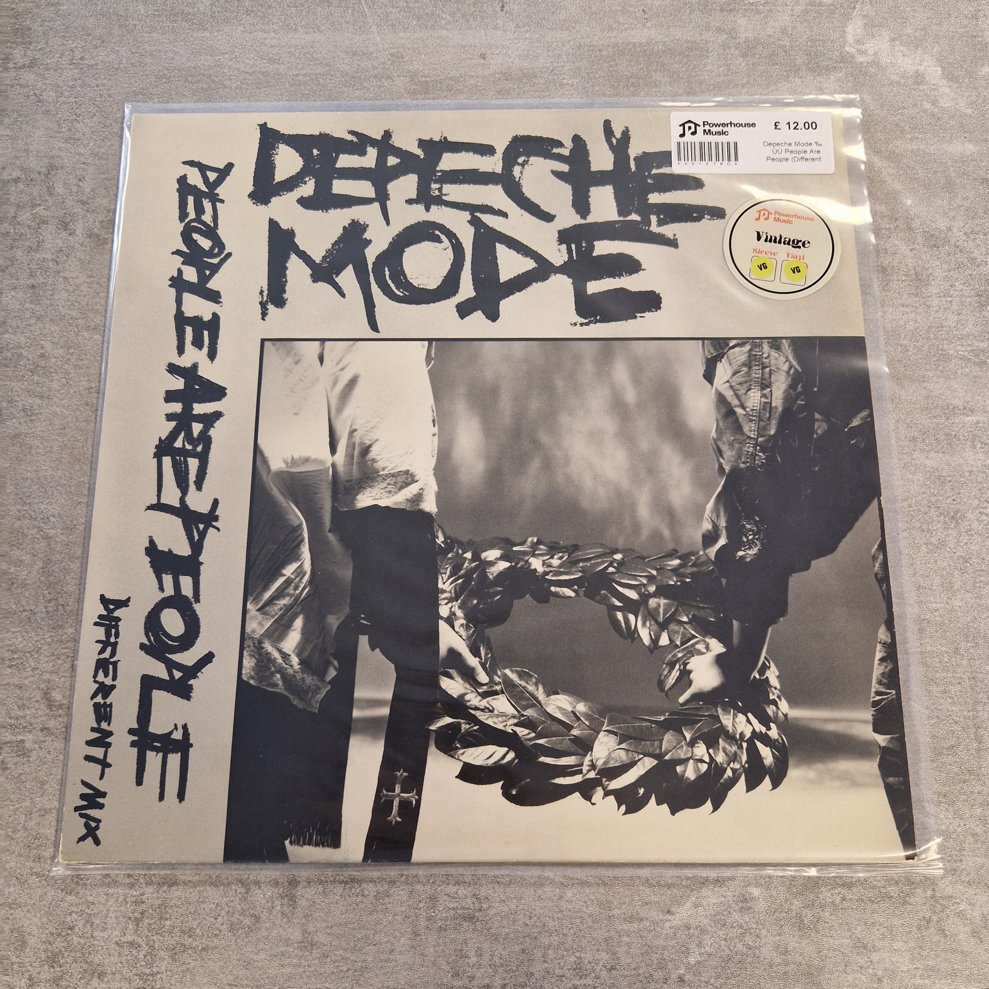 Depeche Mode - People Are People (Different Mix) Vintage Vinyl 12" Single A Depeche Mode vinyl record sleeve in clear plastic shrink-wrap, photographed on a gray textured surface. The cover features the band name 'DEPECHE MODE' in large black brush lettering, 'PEOPLE ARE PEOPLE' vertically on the left, and a black-and-white photo of two people holding a wreath.