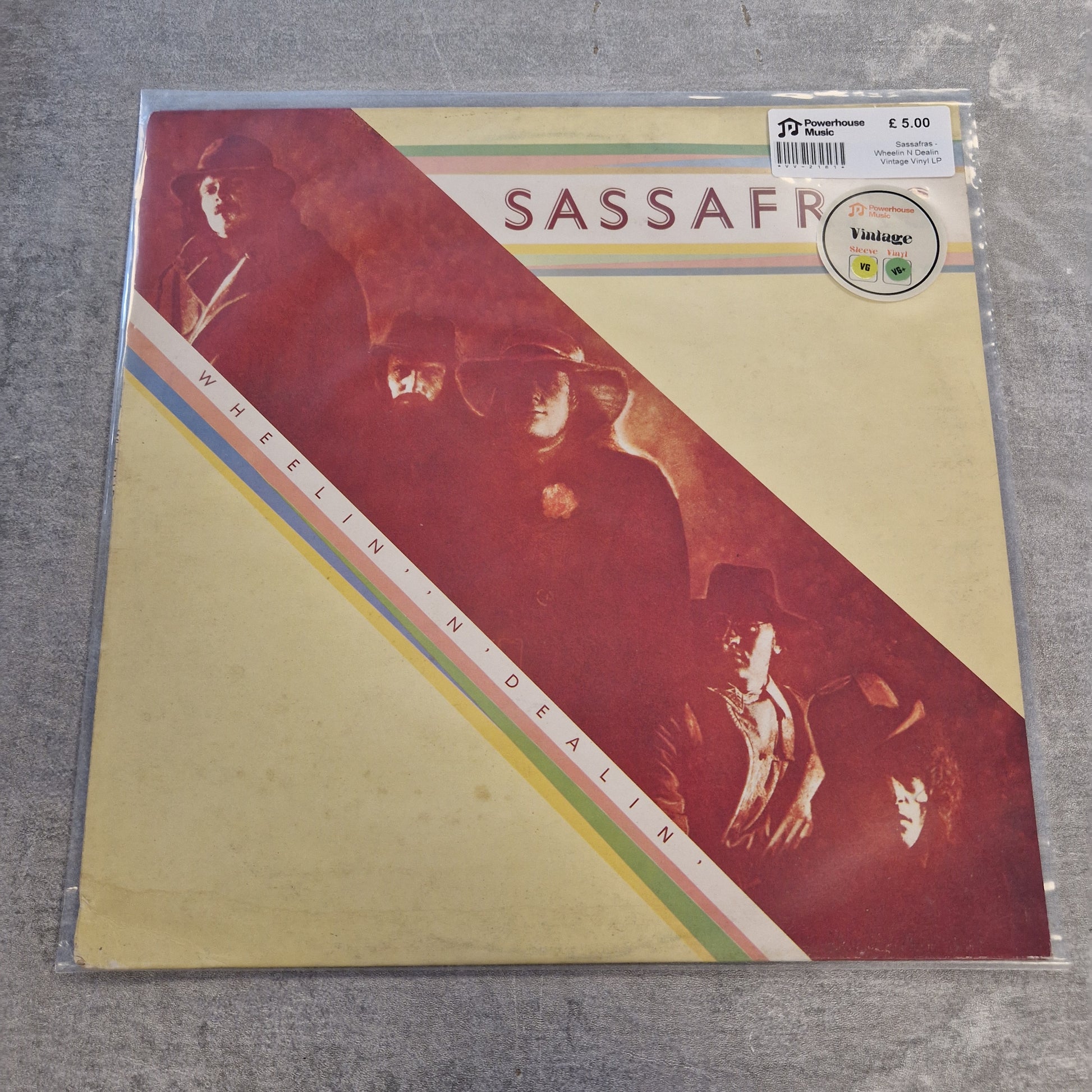 Sassafras - Wheelin N Dealin Vintage Vinyl LP A vinyl record sleeve for Sassafr's 'Shellie N' Dealie N'' on a gray textured surface. The sleeve features a cream-colored background with a large diagonal red graphic, and sepia-toned portraits of the band members.