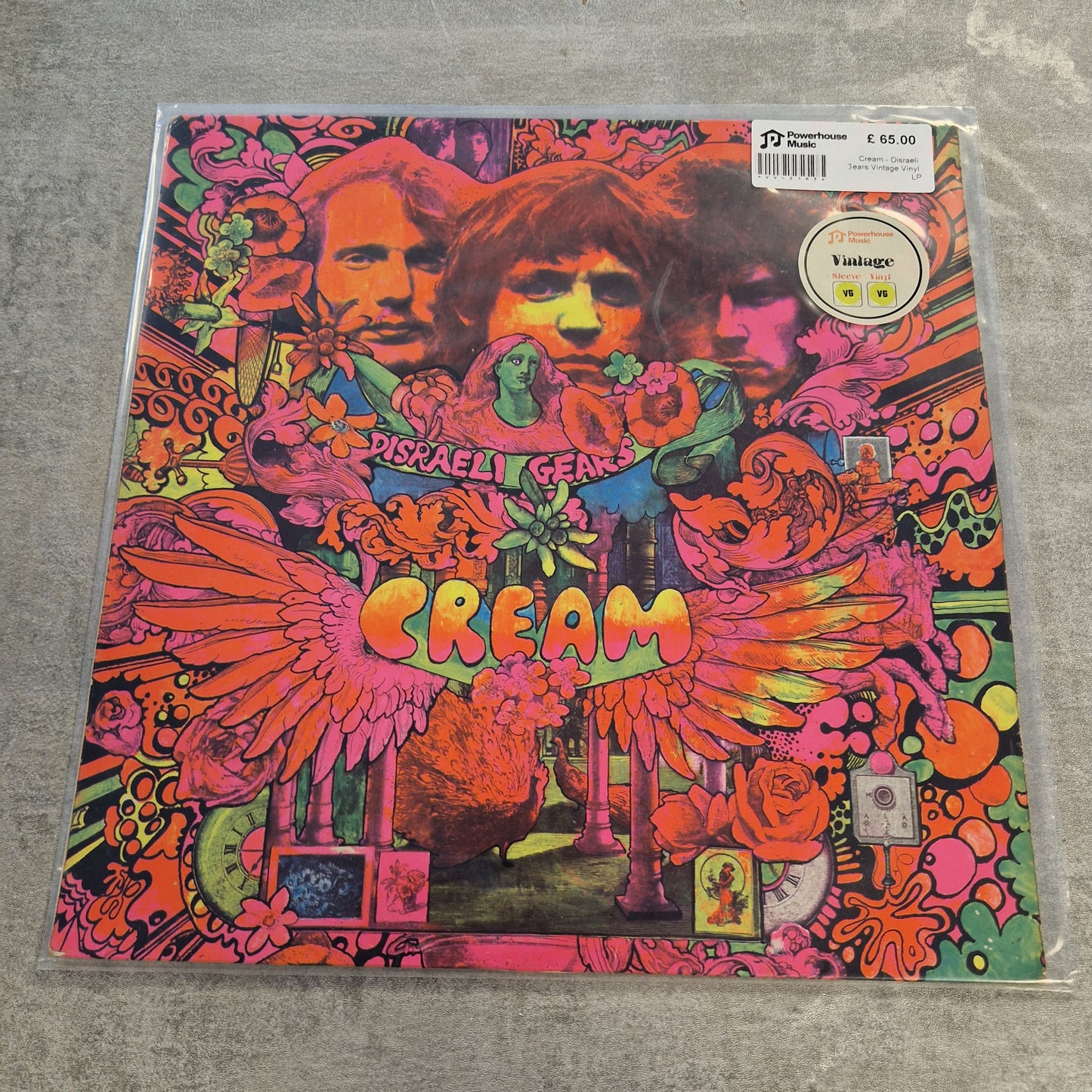 Cream - Disraeli Gears Vintage Vinyl LP A vinyl record of the band Cream's album 'Disraeli Gears' in its clear plastic shrink-wrap, placed on a gray concrete floor. The cover art is highly detailed and psychedelic, featuring the band members' faces, large wings, and the album title in a colorful gradient.
