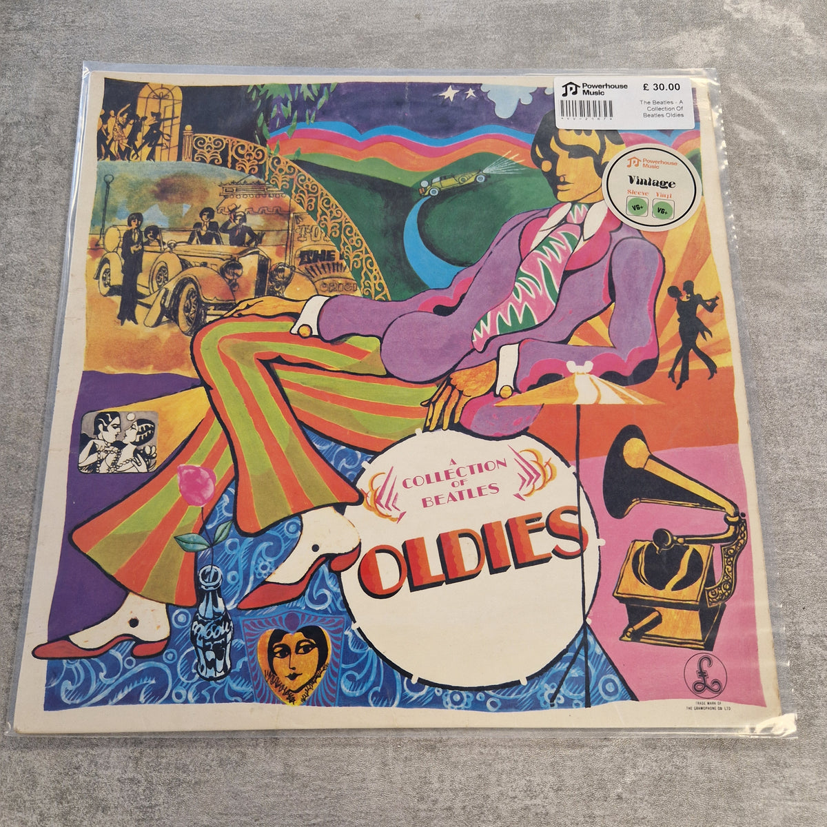 The Beatles - A Collection Of Beatles Oldies Vintage Vinyl LP A vinyl record sleeve for the Beatles' 'Collection of Fabbes Oldies' is shown in its plastic shrink-wrap, lying on a gray textured surface. The cover art is a highly colorful, psychedelic collage featuring a seated figure in a purple suit and green-striped trousers, with a gramophone, car, and rainbow motifs.