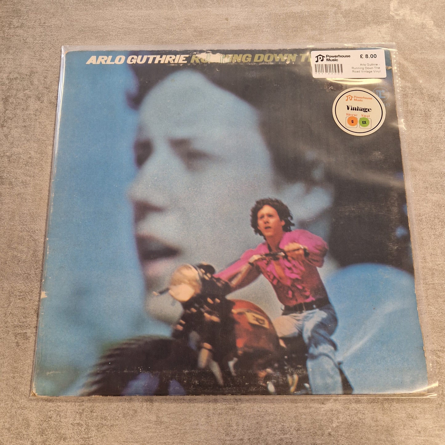Arlo Guthrie - Running Down The Road Vintage Vinyl LP A vinyl record album of Arlo Guthrie's 'Running Down Trough' on the Verve label, photographed on a textured gray surface. The album cover features a large blue-toned portrait of a man in the background with a smaller photo of Arlo Guthrie riding a motorcycle in the foreground.