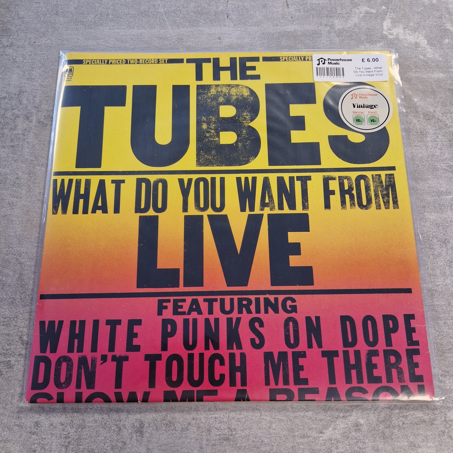 The Tubes - What Do You Want From Live Vintage Vinyl LP The Tubes 'What Do You Want From Live' vinyl record sleeve in its plastic shrink-wrap, displayed on a gray concrete floor. The cover features a bold yellow-to-pink gradient background with black text.