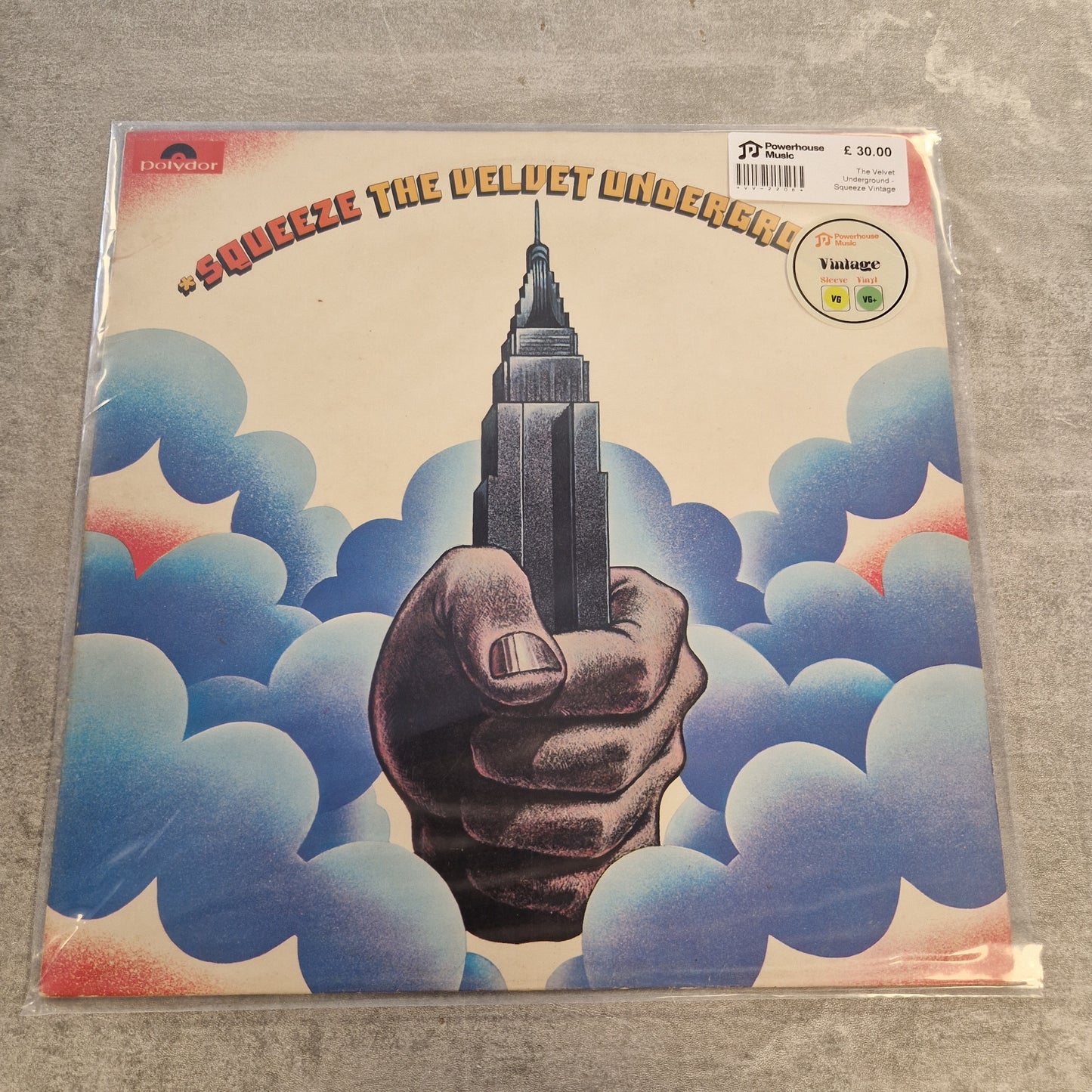 The Velvet Underground - Squeeze Vintage Vinyl LP A vinyl record sleeve for the band One Love and their album 'Squeeze the Velvet Underground' is shown in its plastic shrink-wrap on a gray textured surface. The cover art features a hand holding a pencil shaped like the Empire State Building, surrounded by blue and white clouds with red accents.