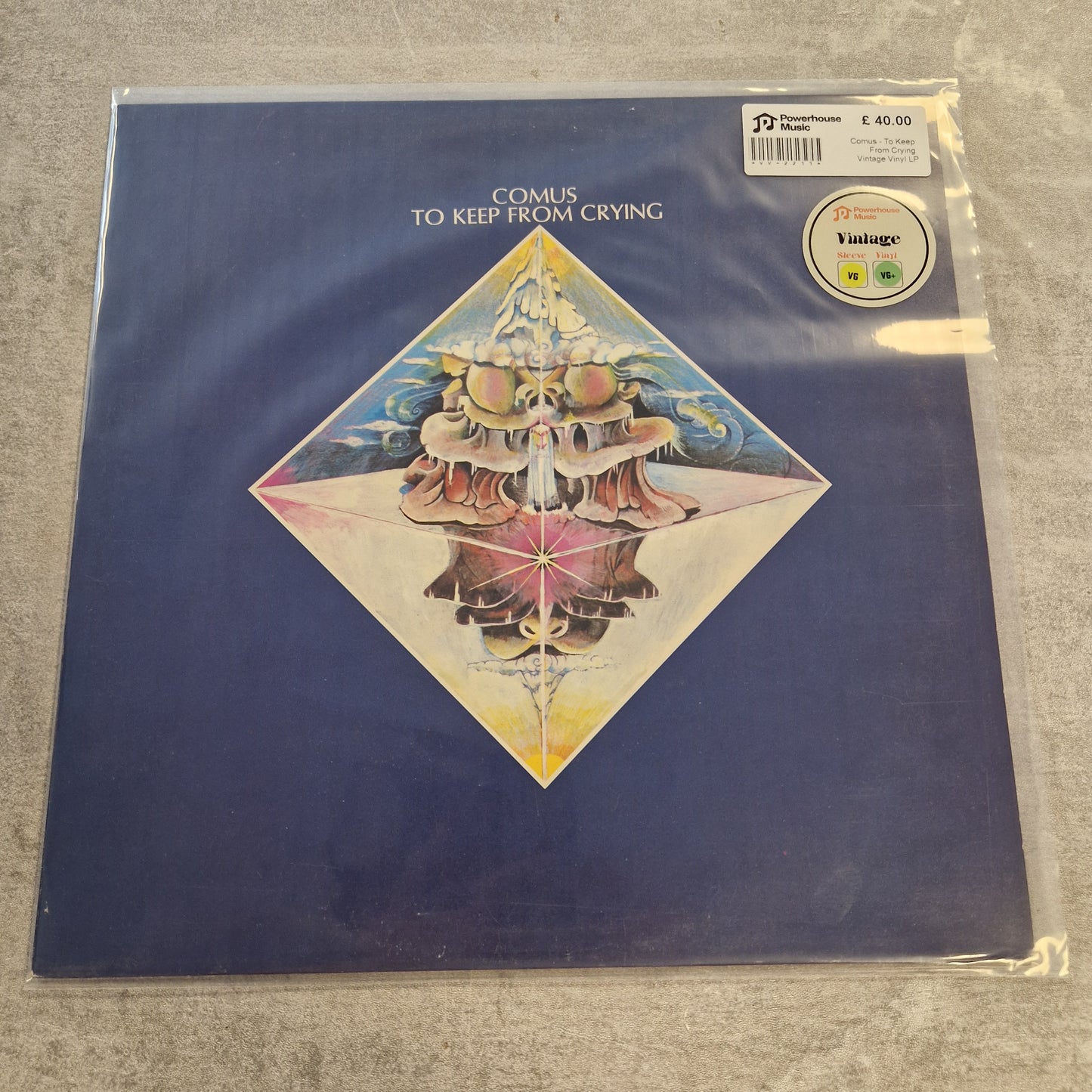 Comus - To Keep From Crying Vintage Vinyl LP A vinyl record sleeve for Comus's 'To Keep from Crying' in a clear plastic protective case, photographed on a gray textured surface. The sleeve features a dark blue background with a central diamond-shaped illustration of a surreal, layered figure in pink, yellow, and blue tones.