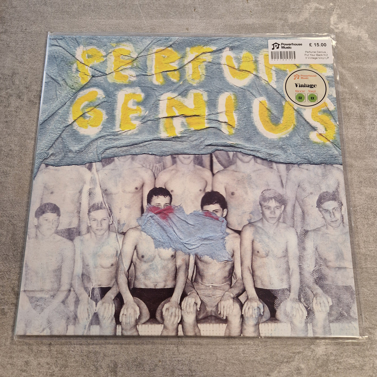 Perfume Genius - Put Your Back N 2 It Vintage Vinyl LP A vinyl record sleeve for the band 'Perfume Genius' lies on a gray concrete floor. The cover art features a blue flag with the band's name in yellow letters above a black-and-white photo of shirtless men.