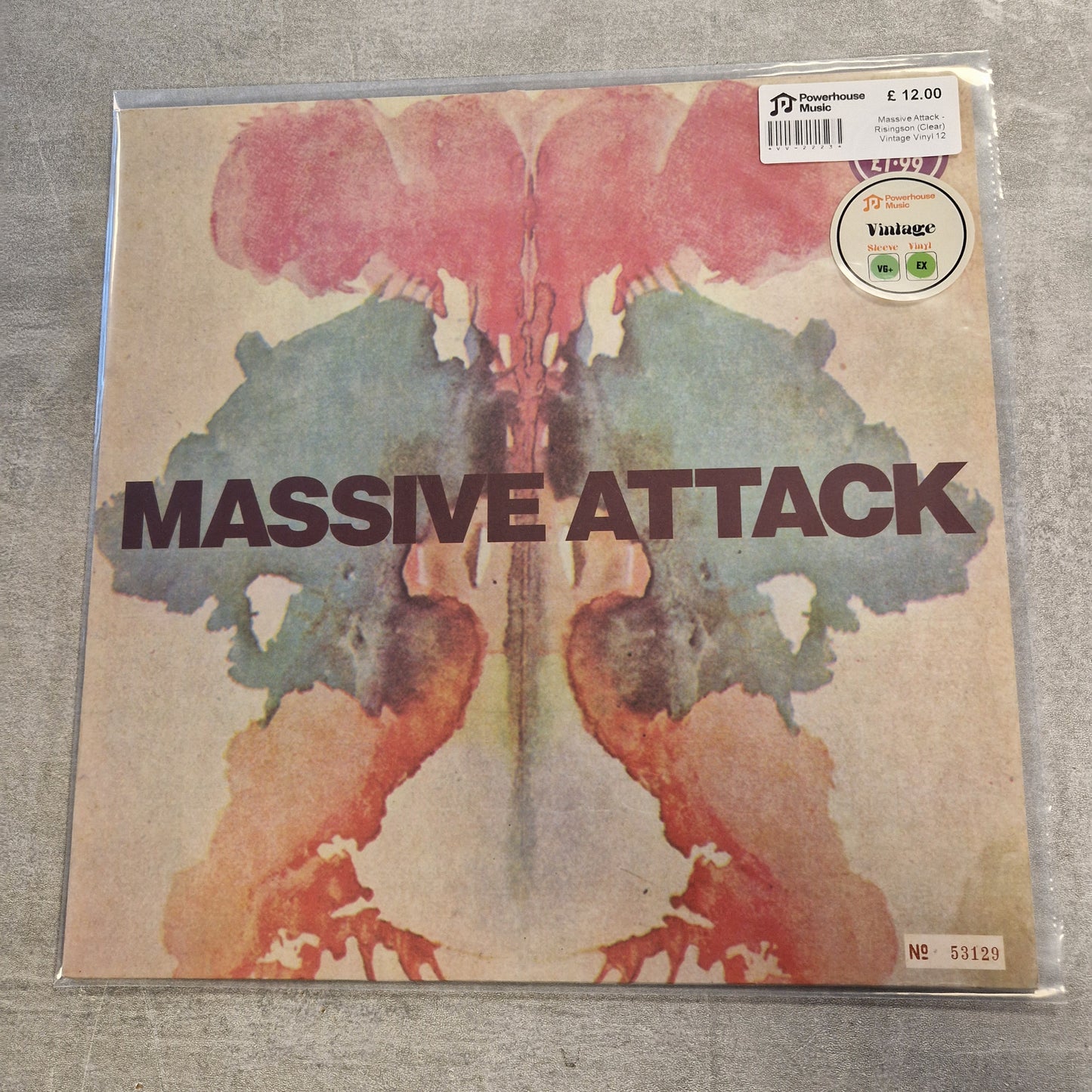 Massive Attack - Risingson (Clear) Vintage Vinyl 12" Single A vinyl record sleeve for Massive Attack's album 'Illside' on a gray textured surface. The cover features a symmetrical, abstract ink-blot design in pink, teal, and orange-brown tones with the band name 'MASSIVE ATTACK' printed across the center.