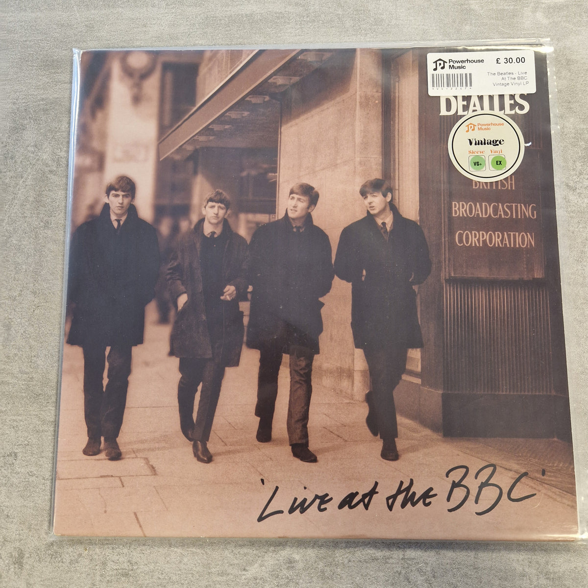 The Beatles - Live At The BBC Vintage Vinyl LP A Beatles 'Live at the BBC' vinyl record sleeve photographed flat on a gray textured surface. The cover shows the band members walking outdoors in dark clothing, with a visible Beatles 'Vintage' sticker and a handwritten signature near the bottom.