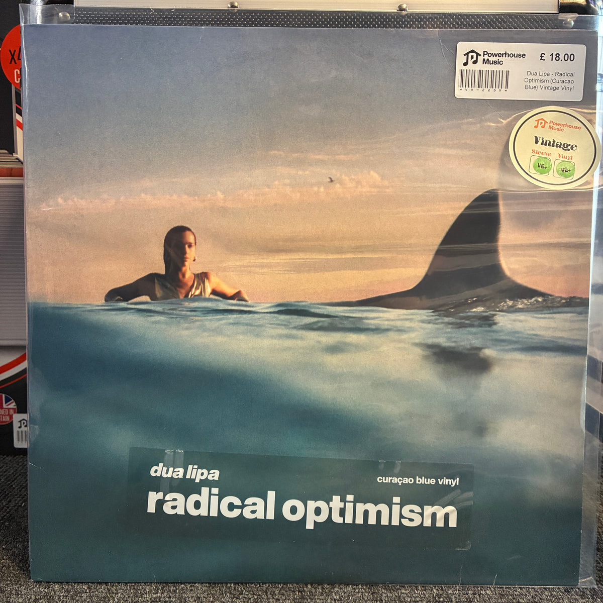Dua Lipa - Radical Optimism (Curacao Blue) Vintage Vinyl LP A vinyl record sleeve for 'radical optimism' by duo lipa, featuring a split water-sky scene with a shark fin and a person in the water. The sleeve is inside a clear protective plastic sleeve, with a Powerhouse label sticker and a Vintage vinyl sticker visible on the top right.