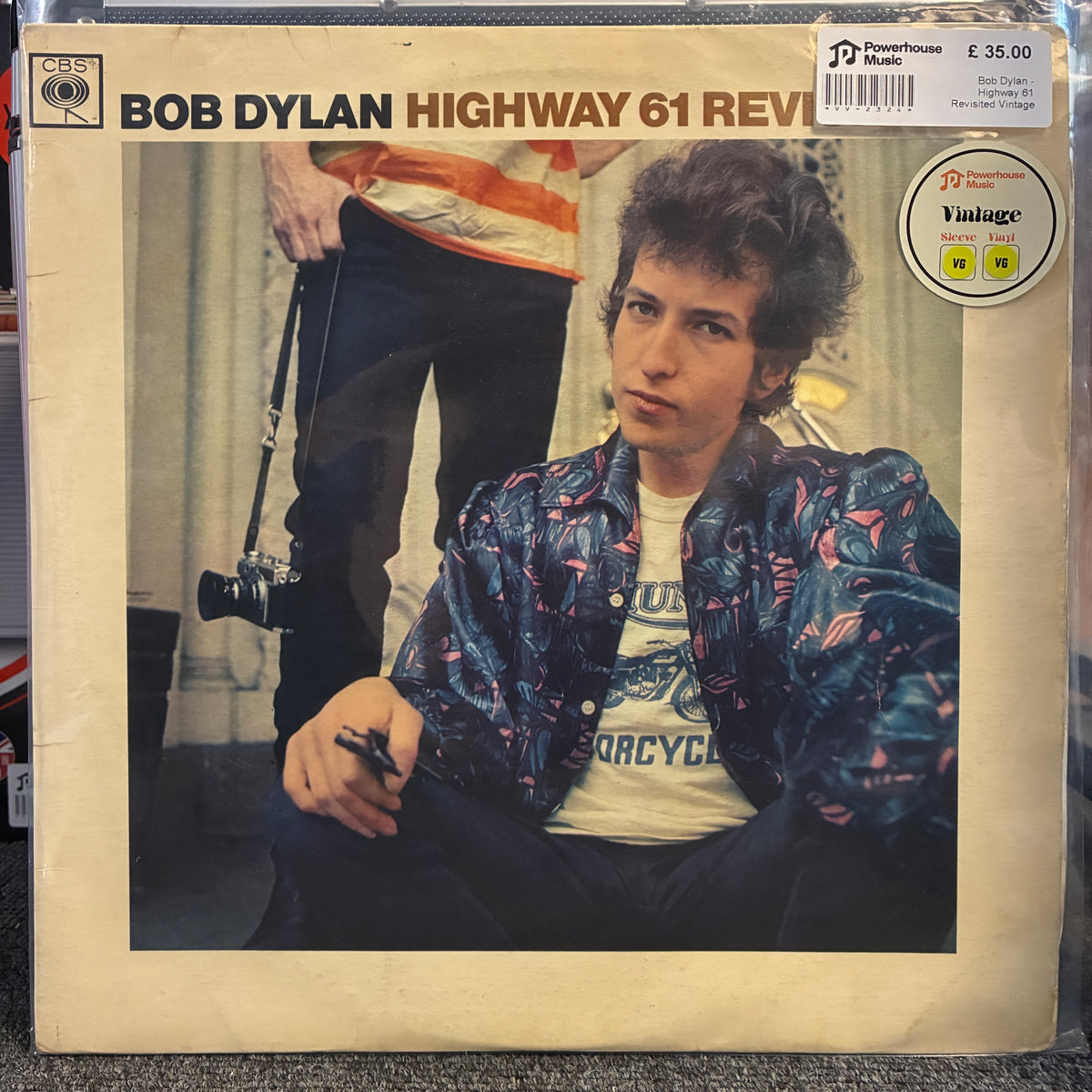 Bob Dylan - Highway 61 Revisited Vintage Vinyl LP Bob Dylan 'Highway 61 Revival' vinyl record album cover, showing a seated Bob Dylan in a patterned jacket and white graphic T-shirt. The cover has a cream border, a barcode sticker at the top right, a 'Vintage Pink' sticker, and a visible 'CBS' logo in the top left.
