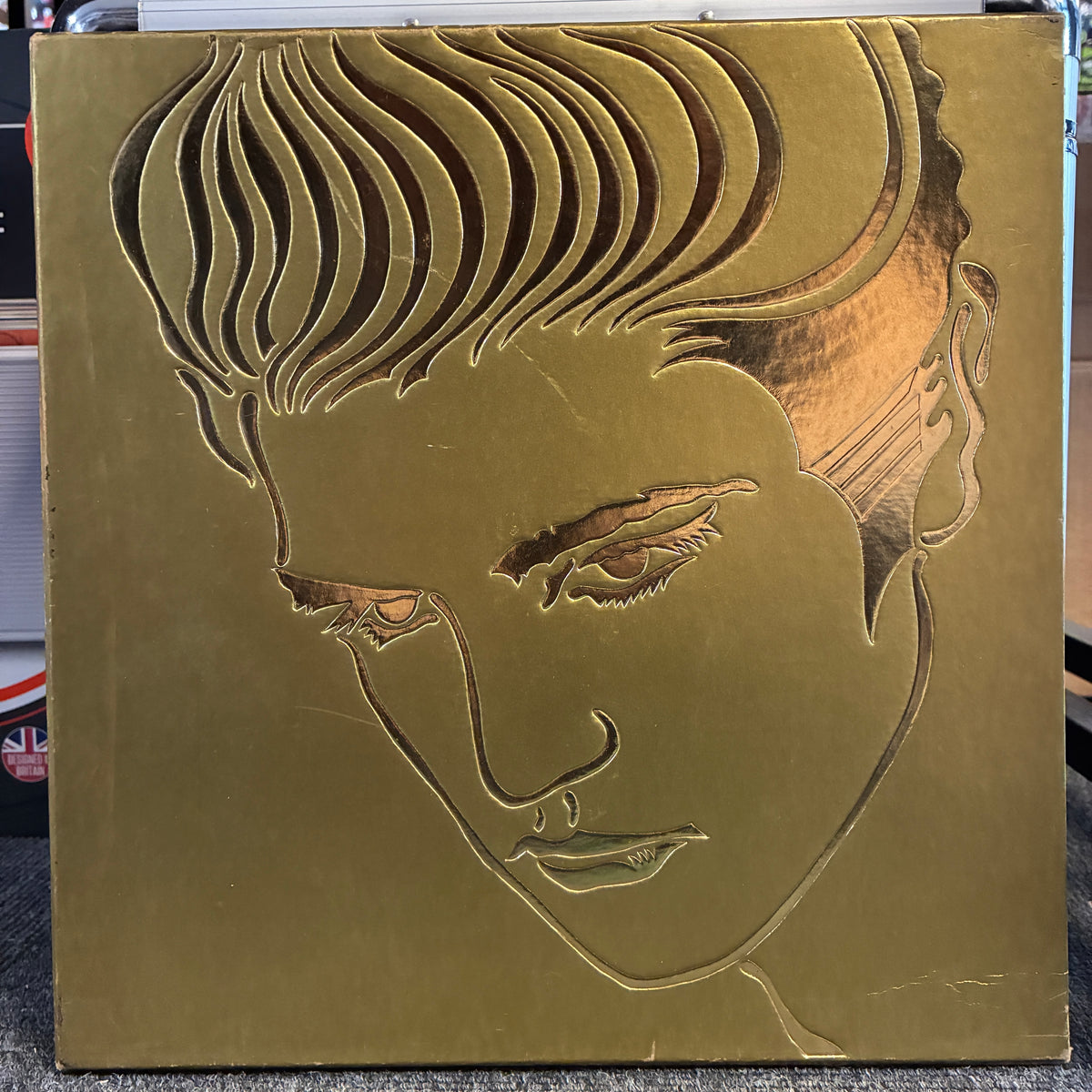 Elvis Presley - A Golden Celebration Vintage Vinyl Boxset A square metallic gold plaque or decorative panel featuring an embossed stylized face with wavy hair, photographed against a dark background.