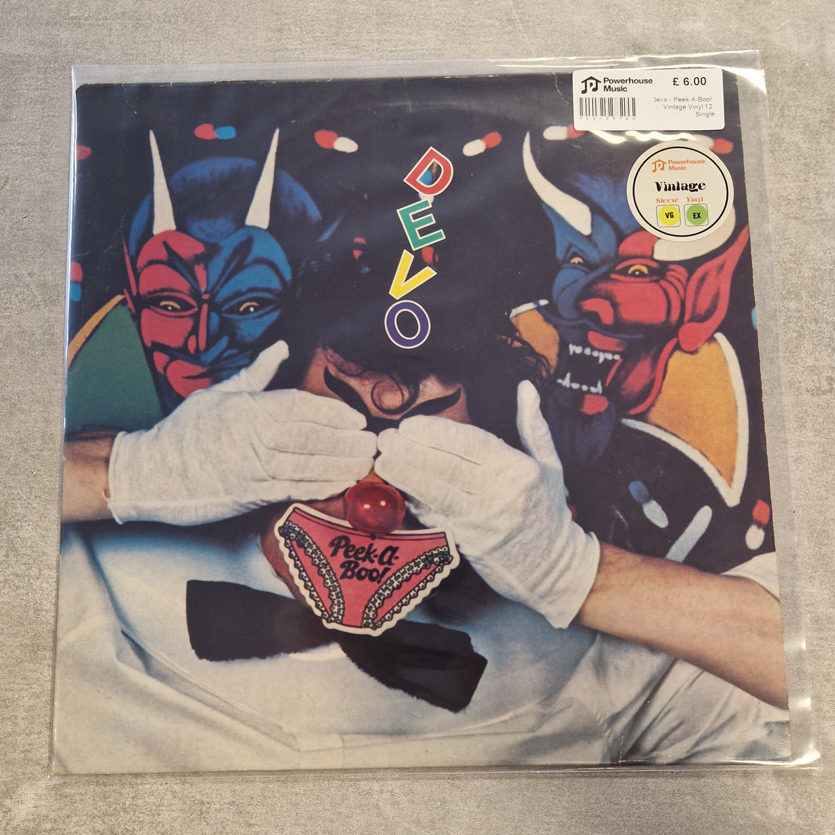 Devo - Peek-A-Boo! Vintage Vinyl 12" Single A vinyl record sleeve for the band DEVO's album 'Park and Recreation' in a clear plastic protective sleeve, photographed on a gray textured surface. The cover art features a dark background with two stylized devil masks (one blue, one red) and a person in white gloves holding a red ball gag, with the word 'DEVO' written vertically in colorful letters.