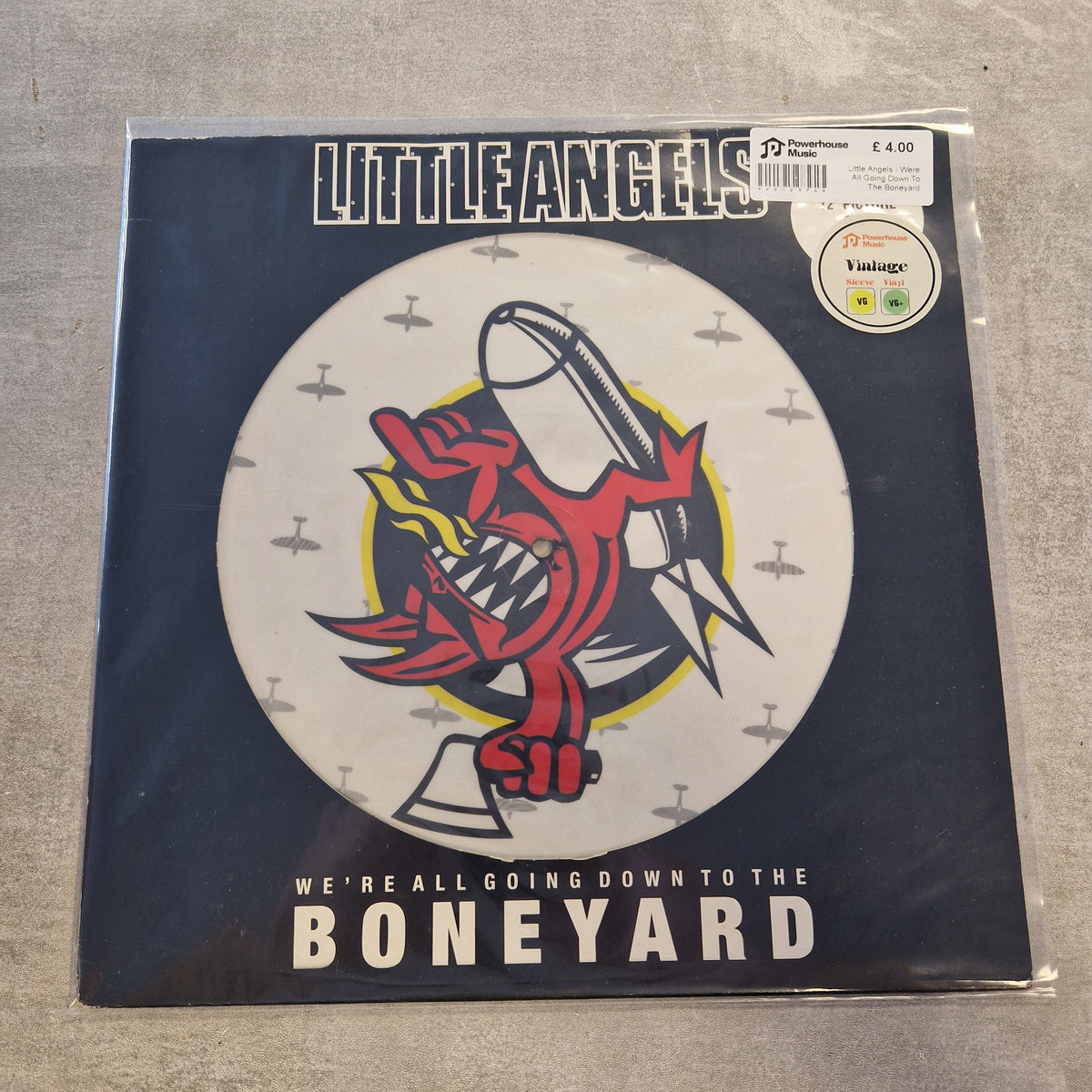 Little Angels - Were All Going Down To The Boneyard (Limited Edition Picture Disc) Vintage Vinyl 12" Single A vinyl record sleeve for Little Angels' 'We're All Going Down to the Boneyard' in a clear plastic protective case, photographed on a gray textured surface. The sleeve features a dark navy blue background with a large white circular graphic containing a red cartoon demon holding a silver bell, with small gray airplane silhouettes.