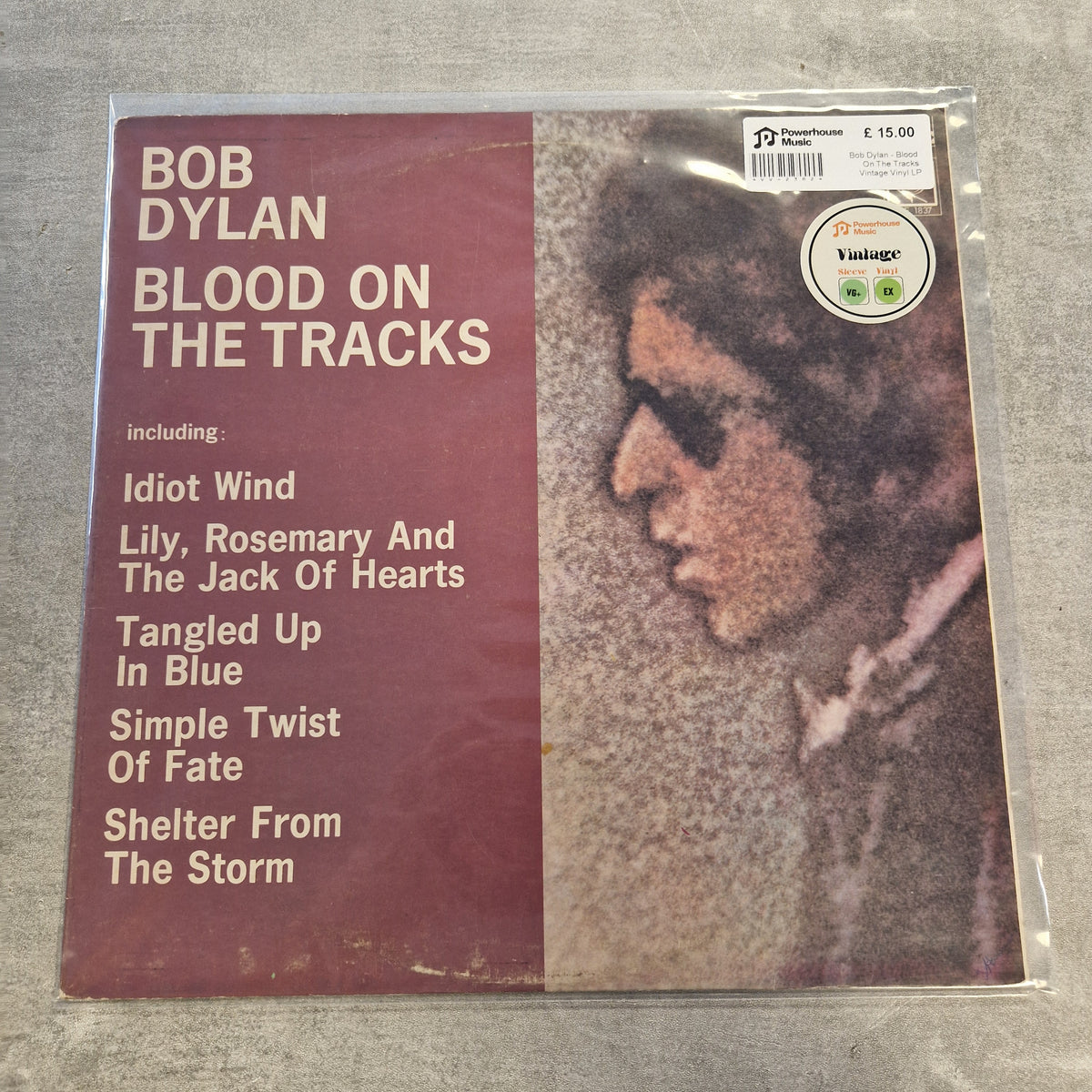 Bob Dylan - Blood On The Tracks Vintage Vinyl LP Bob Dylan 'Blood on the Tracks' vinyl record sleeve in a clear plastic protective case, photographed on a gray textured surface. The cover has a dark red left panel with white text listing track titles and a sepia-toned portrait on the right.