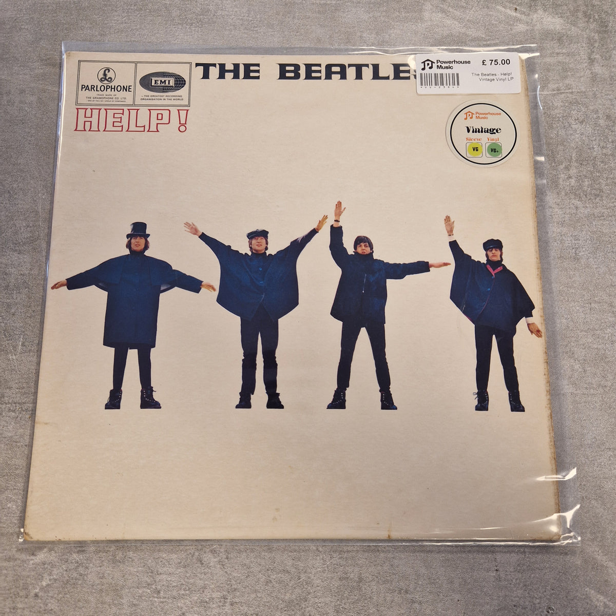 The Beatles - Help! Vintage Vinyl LP The Beatles 'Help!' vinyl record album cover, sealed in a clear plastic sleeve, photographed flat on a gray textured surface. The cover features the band members in blue suits against a cream-colored background, with the Parlophone and EMI logos at the top.