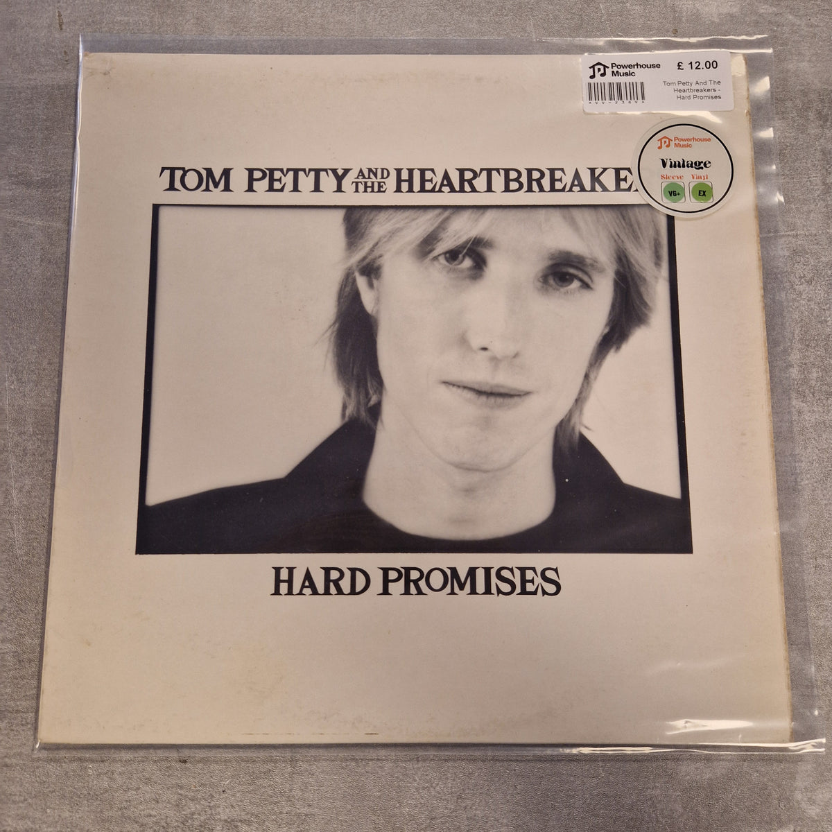 Tom Petty And The Heartbreakers - Hard Promises Vintage Vinyl LP Tom Petty and The Heartbreakers 'Hard Promises' vinyl record sleeve in clear plastic protective wrapping, photographed flat on a gray textured surface. The cover is cream-colored with a black-and-white portrait of Tom Petty and black text.