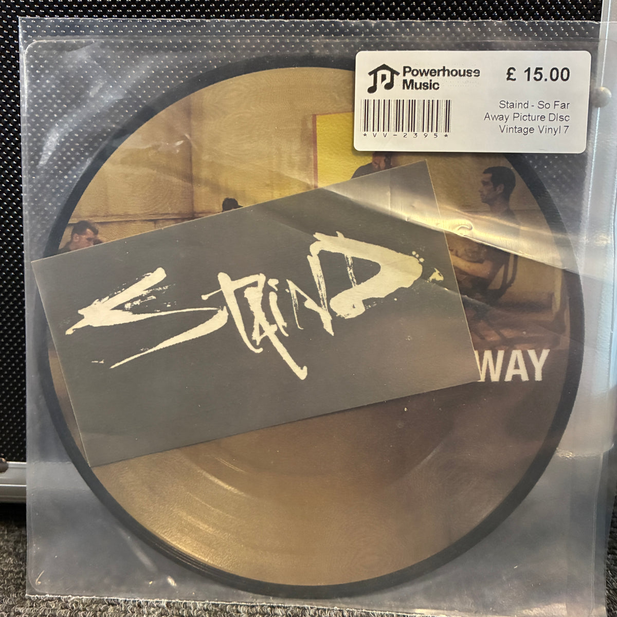 Staind - So Far Away Picture Disc Vintage Vinyl 7" Single A vinyl record package in a clear plastic sleeve, showing a circular gold-toned disc with a large black sticker reading 'Stand - So Far' across the front. A white Powerhouse Music price label is visible at the top right, and part of the album artwork is visible through the plastic.