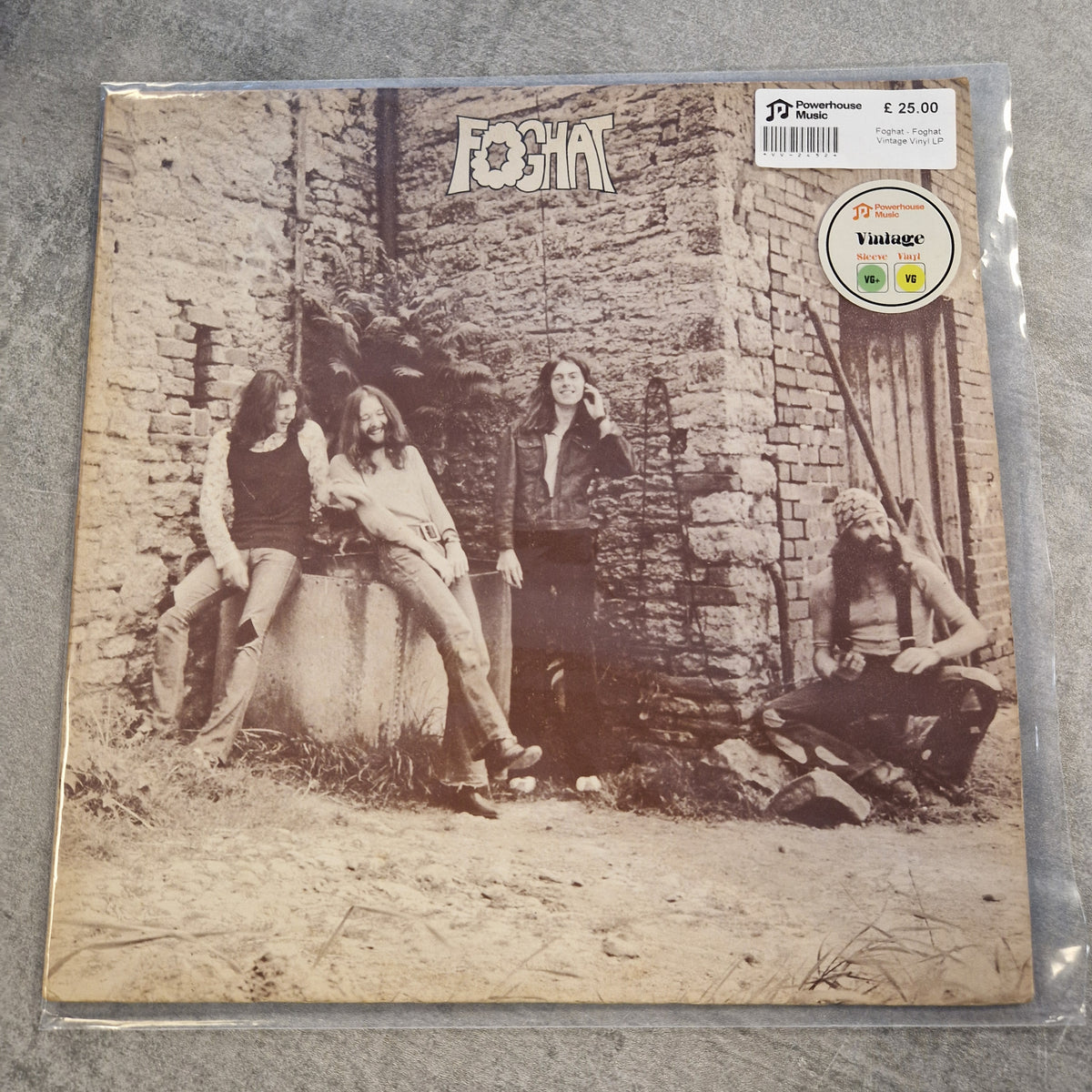 Foghat - Foghat Vintage Vinyl LP A vintage Foghat vinyl record sleeve in clear plastic protective wrapping, placed on a gray textured surface. The square cover features a sepia-toned photograph of four band members in front of a rustic stone wall, with the band name 'FOGCHAT' at the top.