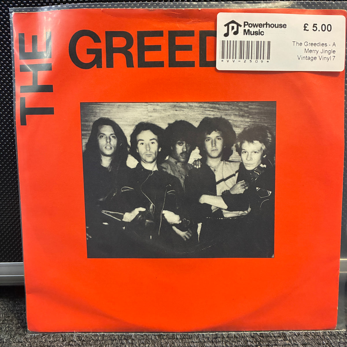 The Greedies - A Merry Jingle Vintage Vinyl 7" Single A vinyl record sleeve for The Greedies' album 'A Merry Jingle' on a bright orange cover, photographed flat against a dark textured background. A white Powerhouse Music sticker is visible in the top right corner.