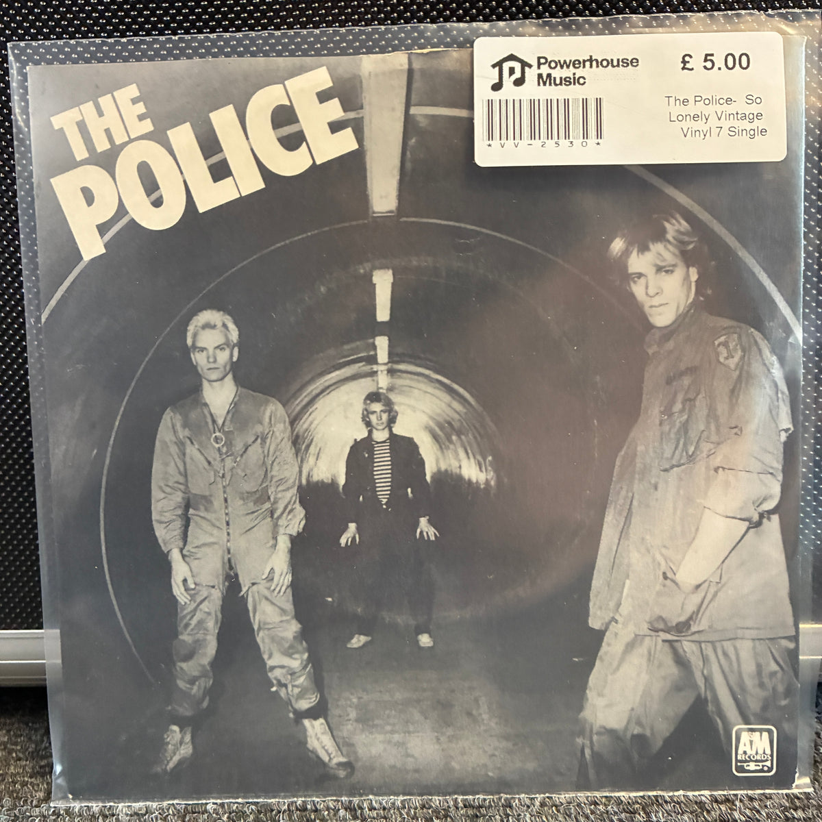 The Police - So Lonely Vintage Vinyl 7" Single The Police 'So Lonely Vintage' vinyl single jacket in a clear plastic sleeve, photographed against a dark textured background. A white barcode sticker for 'Powerhouse Music' is visible in the top right corner.