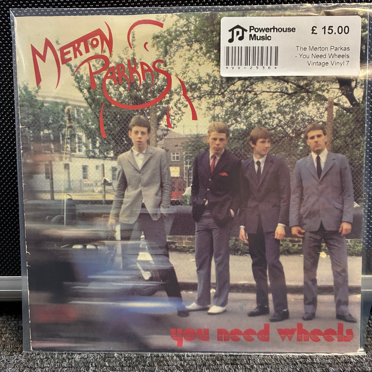 The Merton Parkas - You Need Wheels Vintage Vinyl 7" Single Merton Parkas 'You Need Wheels' vintage vinyl record sleeve on a clear plastic jewel case, displayed on a dark textured surface. The cover features the band members in suits against a blurred street background with a red stylized logo and the title in red text.