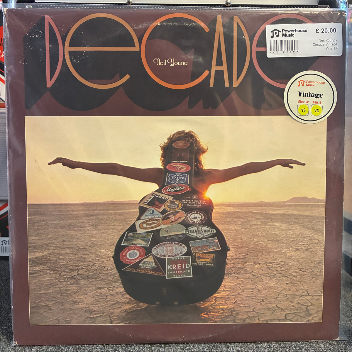 Neil Young - Decade Vintage Vinyl LP Neil Young 'Decade' vinyl record album cover, showing a person standing in a desert landscape with arms outstretched, wearing a guitar-shaped outfit covered in stickers. The cover has a brown border and a 'Vintage' sticker on the top right.