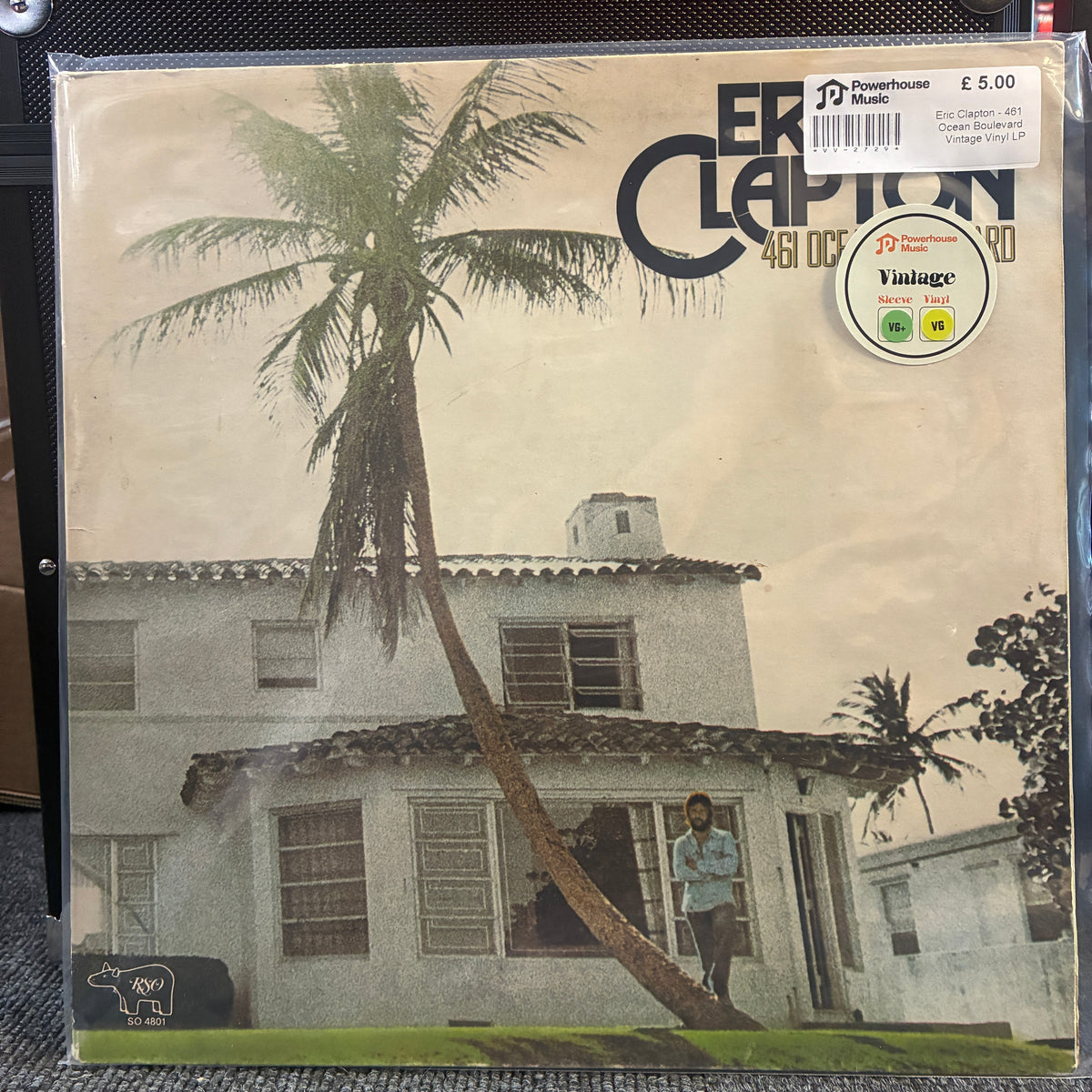 Eric Clapton - 461 Ocean Boulevard Vintage Vinyl LP A vintage vinyl record sleeve for Eric Clapton's '451 Degrees of Freedom' is shown in a clear plastic protective case. The cover art features a sepia-toned photograph of a house with a leaning palm tree and a figure standing outside, with Eric Clapton's name printed in large black letters at the top right.