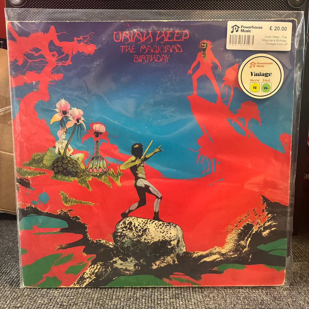Uriah Heep - The Magician's Birthday Vintage Vinyl LP A vinyl record sleeve for Urynh Deep's 'The Psychics Birthday' is shown in a clear plastic protective case on a gray carpet. The cover art features a surreal landscape with red rock formations, a blue sky, and stylized figures.