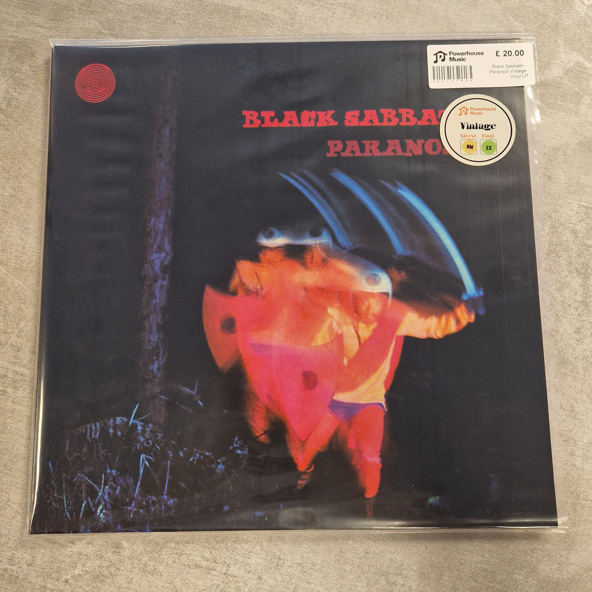 Black Sabbath - Paranoid Vintage Vinyl LP A vintage Black Sabbath 'Paranoic' vinyl record sleeve in clear plastic protective packaging, placed on a gray textured surface. The sleeve features the band's name in red text and an abstract, blurred graphic of figures in red, orange, and blue tones against a dark background.