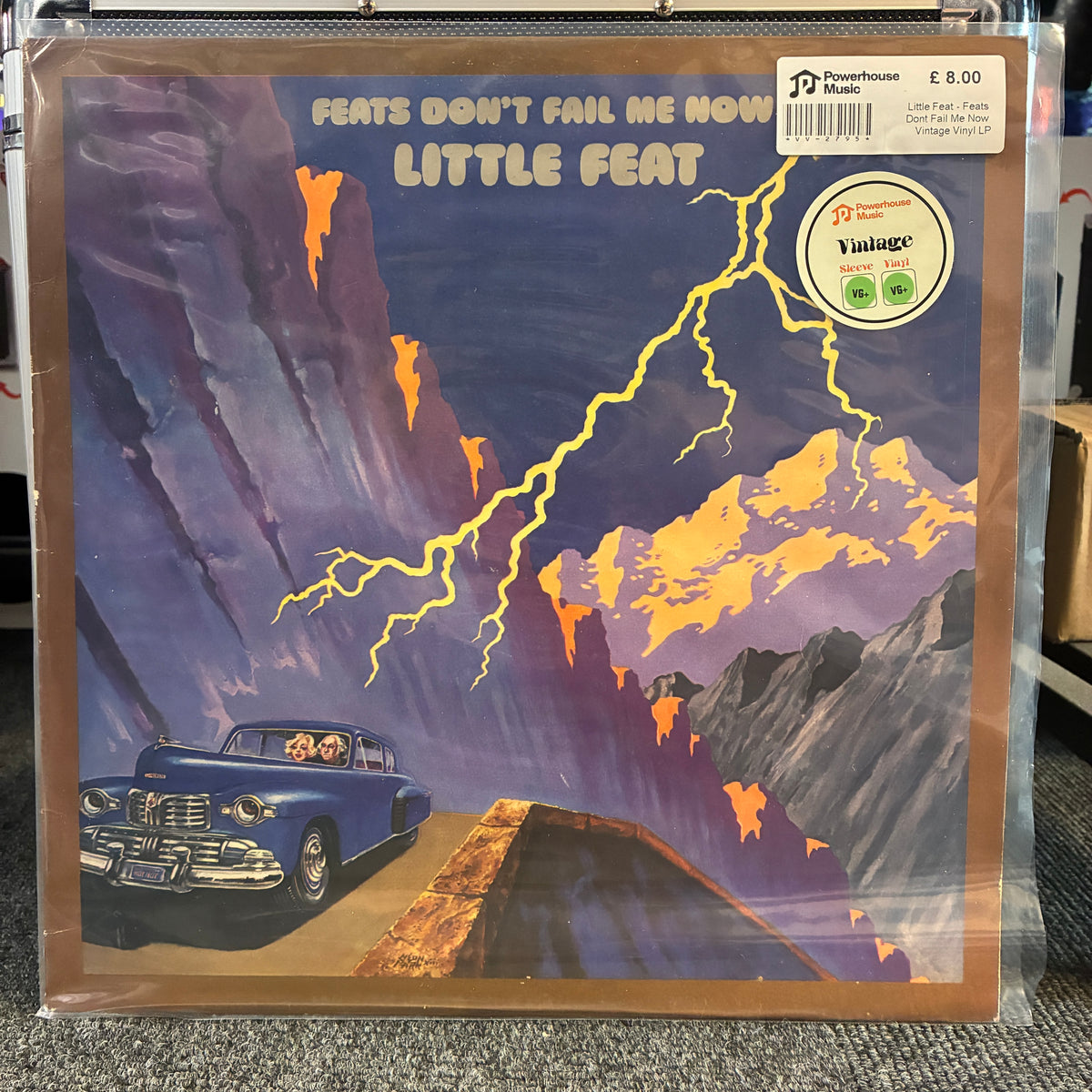 Little Feat - Feats Don't Fail Me Now Vintage Vinyl LP A vinyl record sleeve for Little Feat's 'Feats Don't Fail Me Now' is shown in plastic shrink-wrap, with a barcode sticker and a Vintage logo sticker on the cover. The artwork depicts a blue convertible car driving along a road through a lightning-ridden mountain landscape.