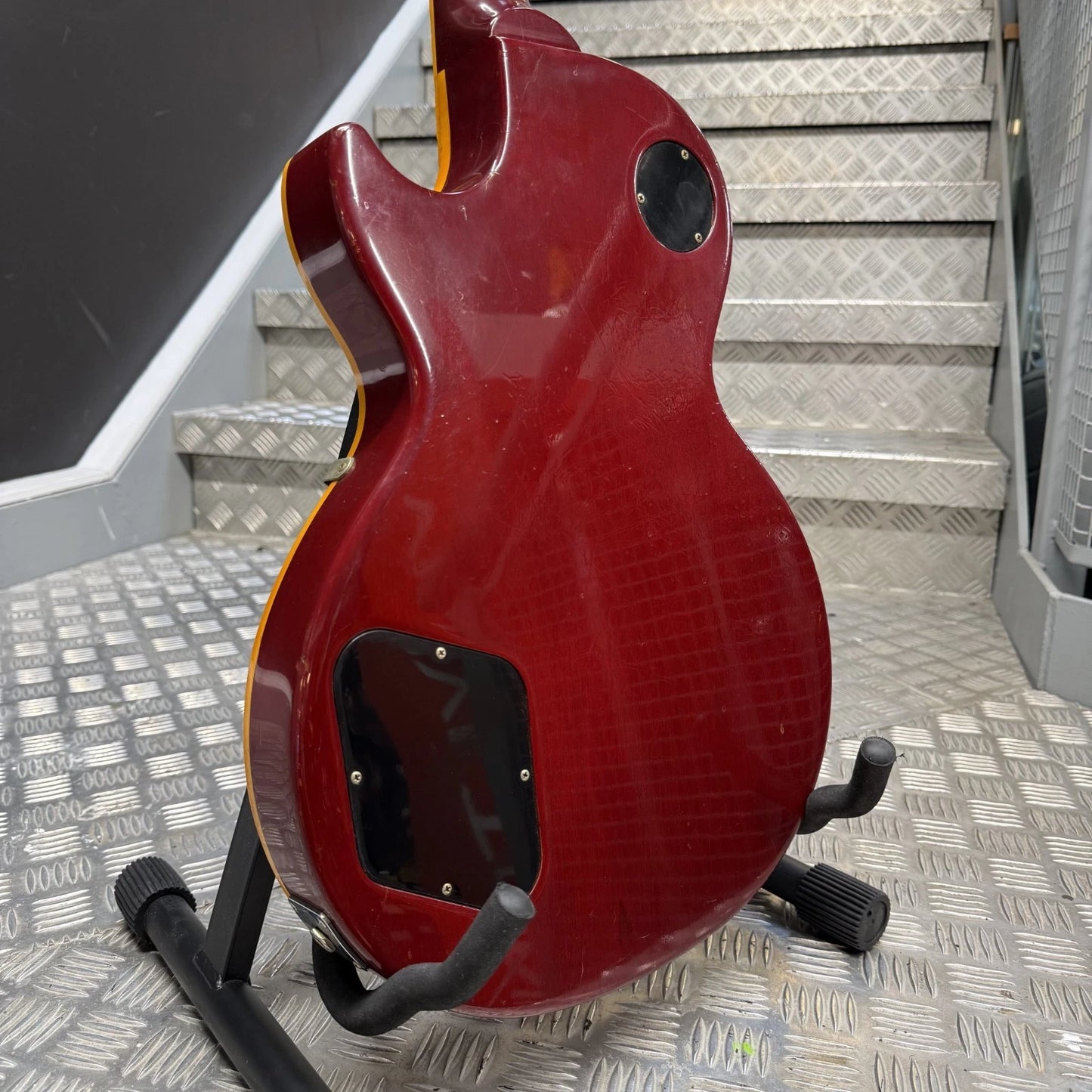 Pre-Owned Gibson Les Paul Classic '1960' 2001 Electric - Cherry Sunburst A red electric guitar body, likely a Gibson Les Paul style, displayed on a black stand on a diamond-plate metal staircase.
