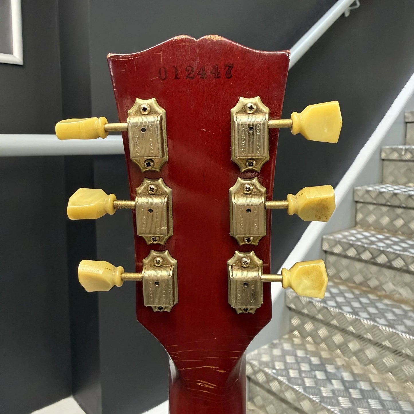 Pre-Owned Gibson Les Paul Classic '1960' 2001 Electric - Cherry Sunburst Close-up of the headstock of a red electric guitar, showing six gold tuning pegs and serial number 012437, with metal stairs in the background.