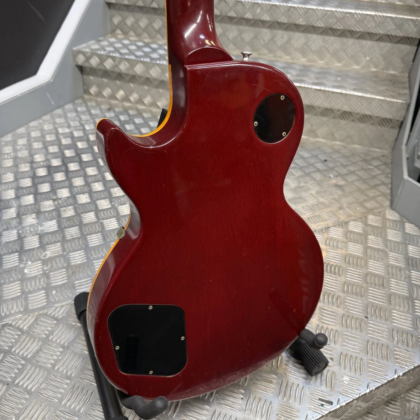 Pre-Owned Gibson Les Paul Classic '1960' 2001 Electric - Cherry Sunburst The rear view of a Gibson Les Paul Custom electric guitar in a glossy dark red finish, resting on a black guitar stand on a diamond plate metal surface.