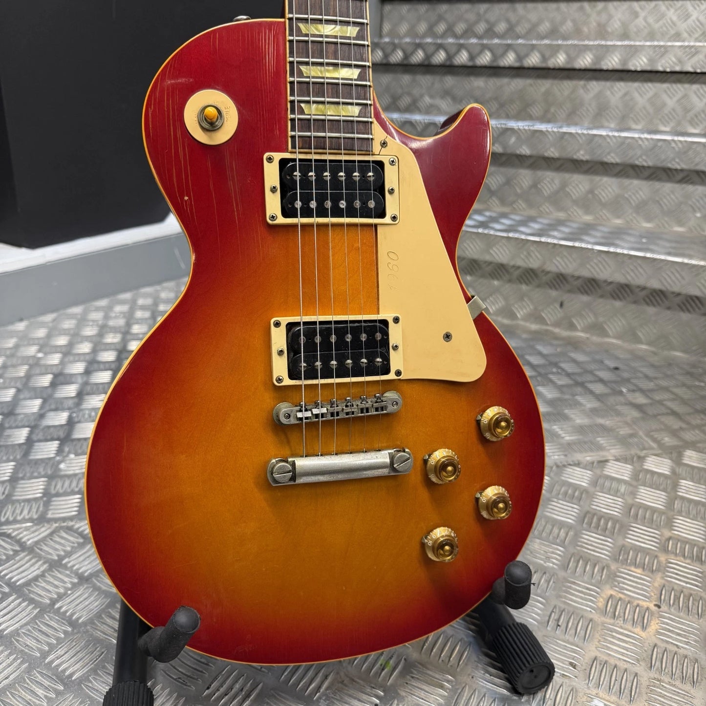 Pre-Owned Gibson Les Paul Classic '1960' 2001 Electric - Cherry Sunburst A Gibson Les Paul Custom electric guitar with a sunburst finish, resting on a black stand against a textured metal background.