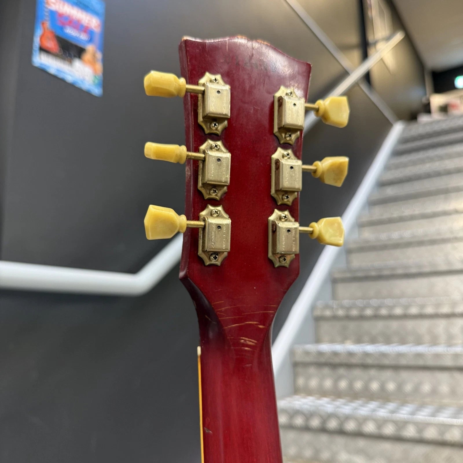 Pre-Owned Gibson Les Paul Classic '1960' 2001 Electric - Cherry Sunburst Close-up of the headstock of a red acoustic guitar, showing six gold tuning pegs, with a blurred staircase in the background.