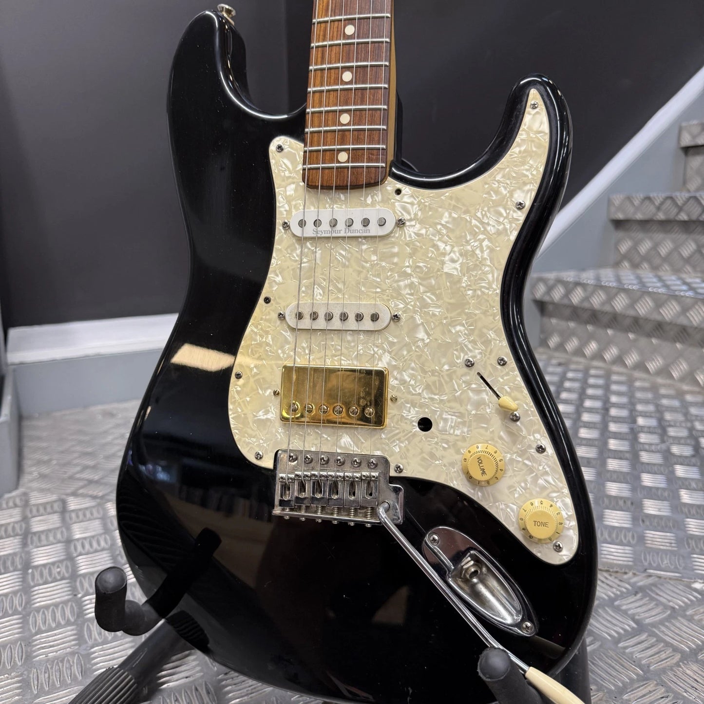 Pre-Owned Fender USA American Traditional Fat Stratocaster (Export Model) 1999 Electric - Black A black electric guitar with a cream pearlescent pickguard and gold hardware, shown resting on a stand in front of metal stairs.