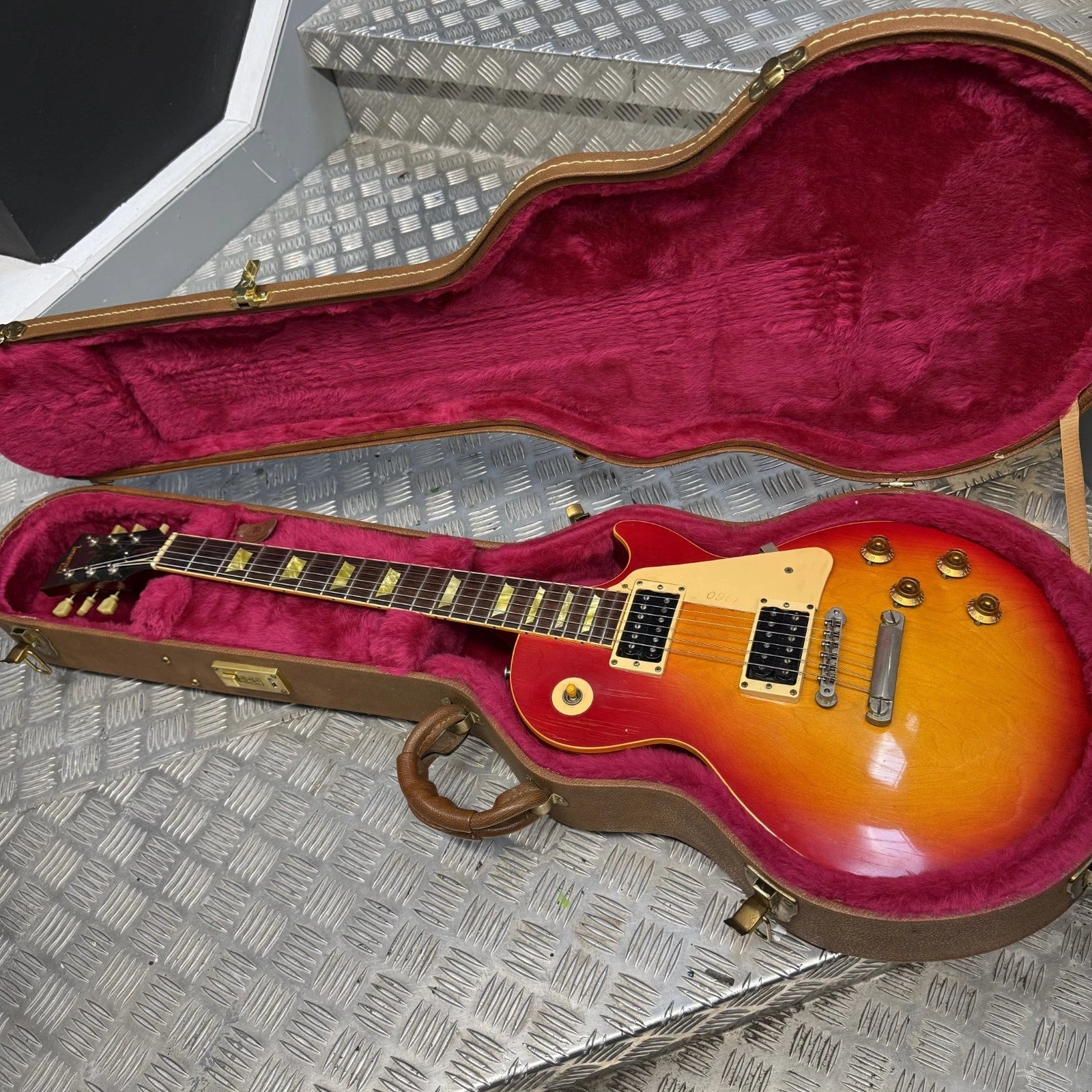 Pre-Owned Gibson Les Paul Classic '1960' 2001 Electric - Cherry Sunburst An open Gibson electric guitar case with a brown hard shell and deep magenta velvet lining, containing a sunburst electric guitar on a diamond plate metal surface.