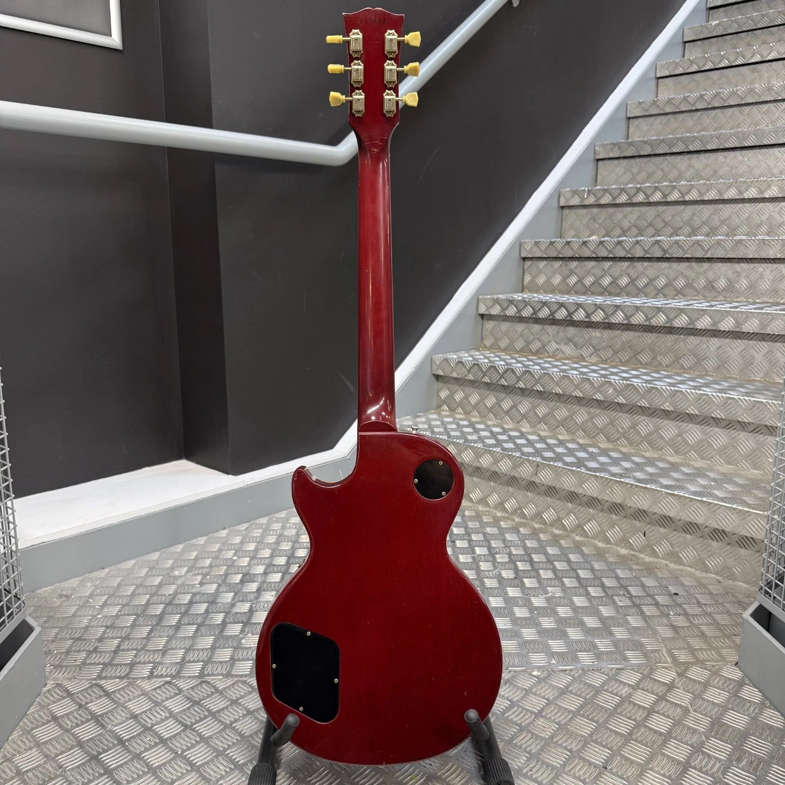 Pre-Owned Gibson Les Paul Classic '1960' 2001 Electric - Cherry Sunburst A red electric guitar shown from the back, standing on a stand in front of a staircase with diamond-plate steps and a dark gray wall.