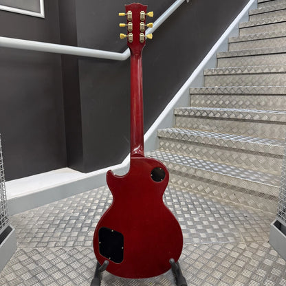 Pre-Owned Gibson Les Paul Classic '1960' 2001 Electric - Cherry Sunburst A red electric guitar shown from the back, standing on a stand in front of a staircase with diamond-plate steps and a dark gray wall.