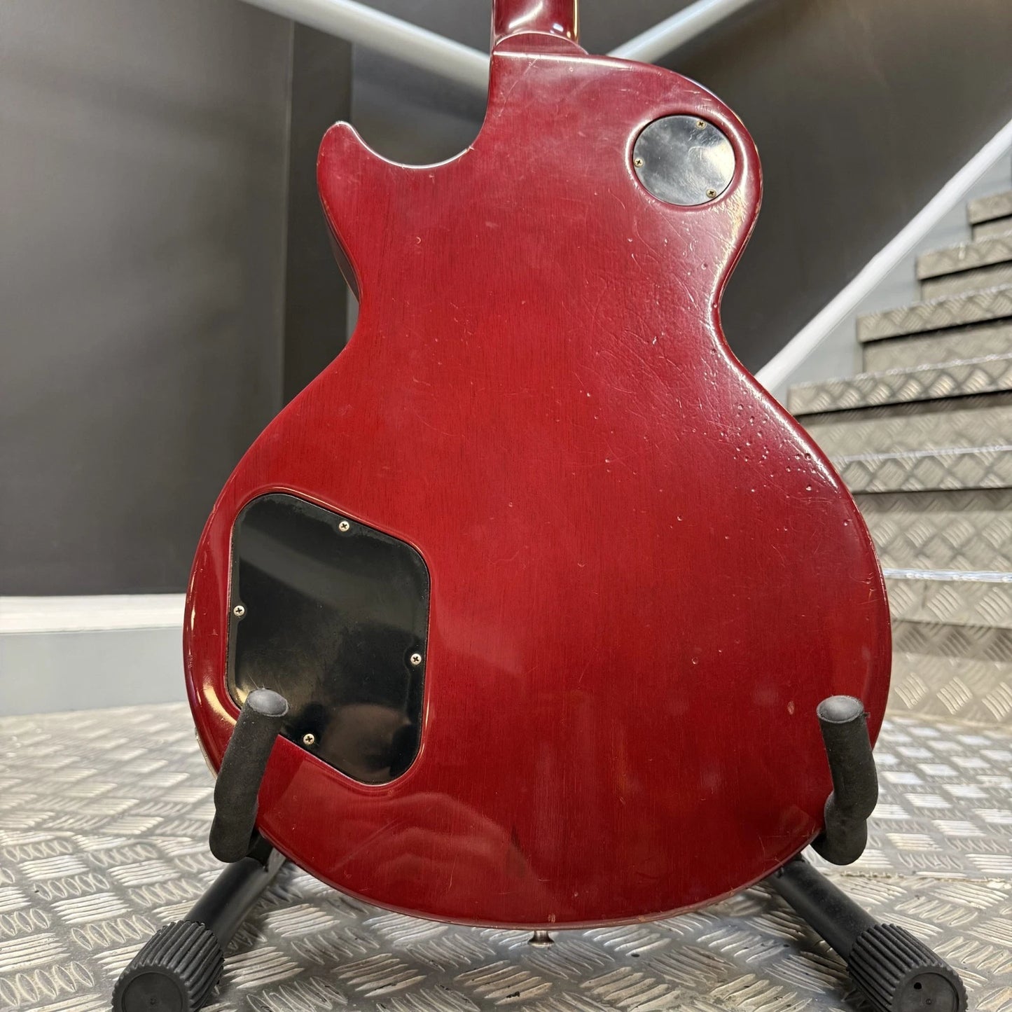 Pre-Owned Gibson Les Paul Classic '1960' 2001 Electric - Cherry Sunburst The back of a red electric guitar on a black stand, showing a glossy red finish, a circular silver control, and a black rectangular plate cutout, set against a background of gray metal steps and a dark wall.