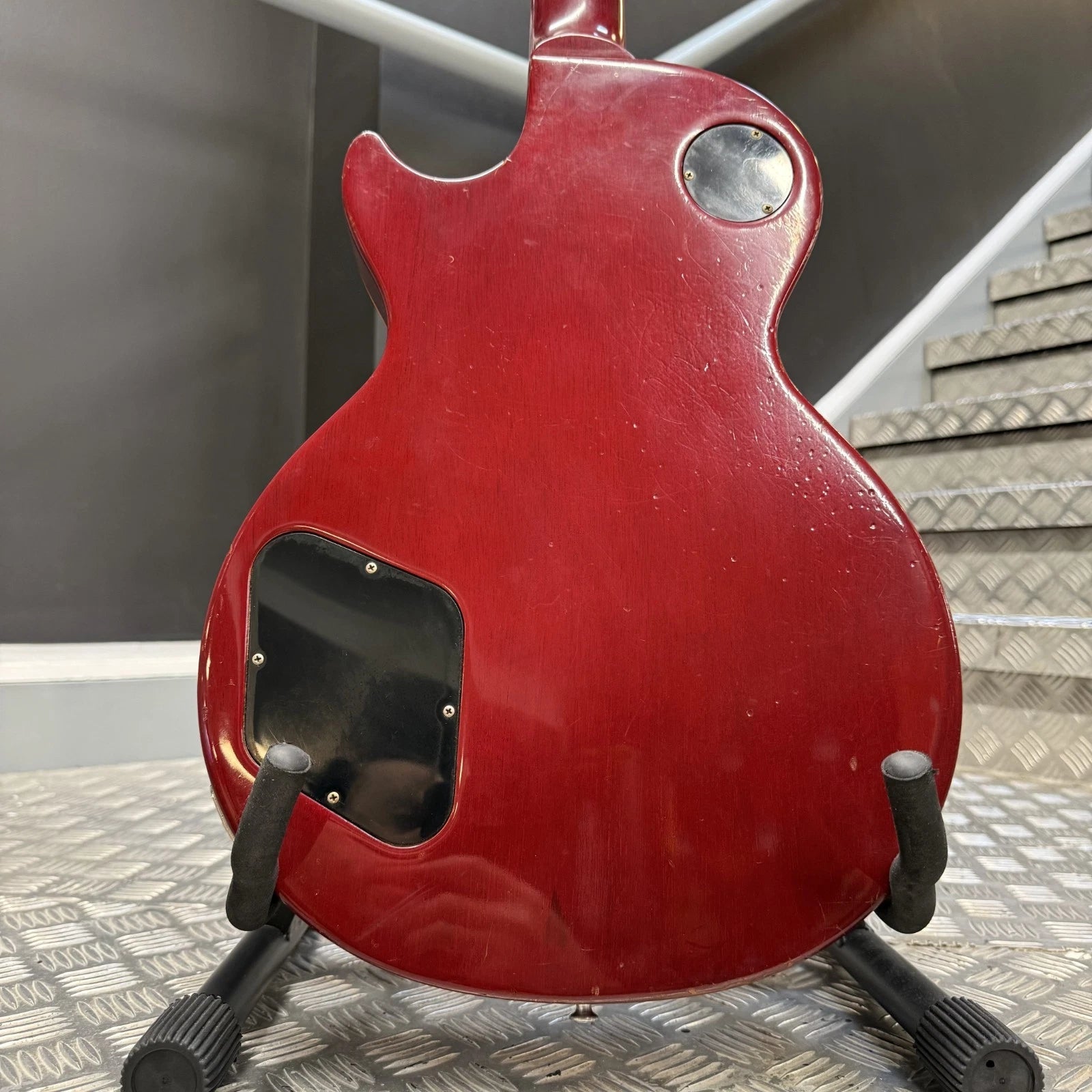 Pre-Owned Gibson Les Paul Classic '1960' 2001 Electric - Cherry Sunburst The back of a red electric guitar on a black stand, showing a glossy red finish, a circular silver control, and a black rectangular plate cutout, set against a background of gray metal steps and a dark wall.