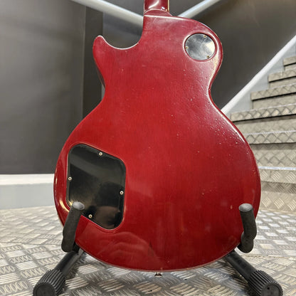 Pre-Owned Gibson Les Paul Classic '1960' 2001 Electric - Cherry Sunburst The back of a red electric guitar on a black stand, showing a glossy red finish, a circular silver control, and a black rectangular plate cutout, set against a background of gray metal steps and a dark wall.