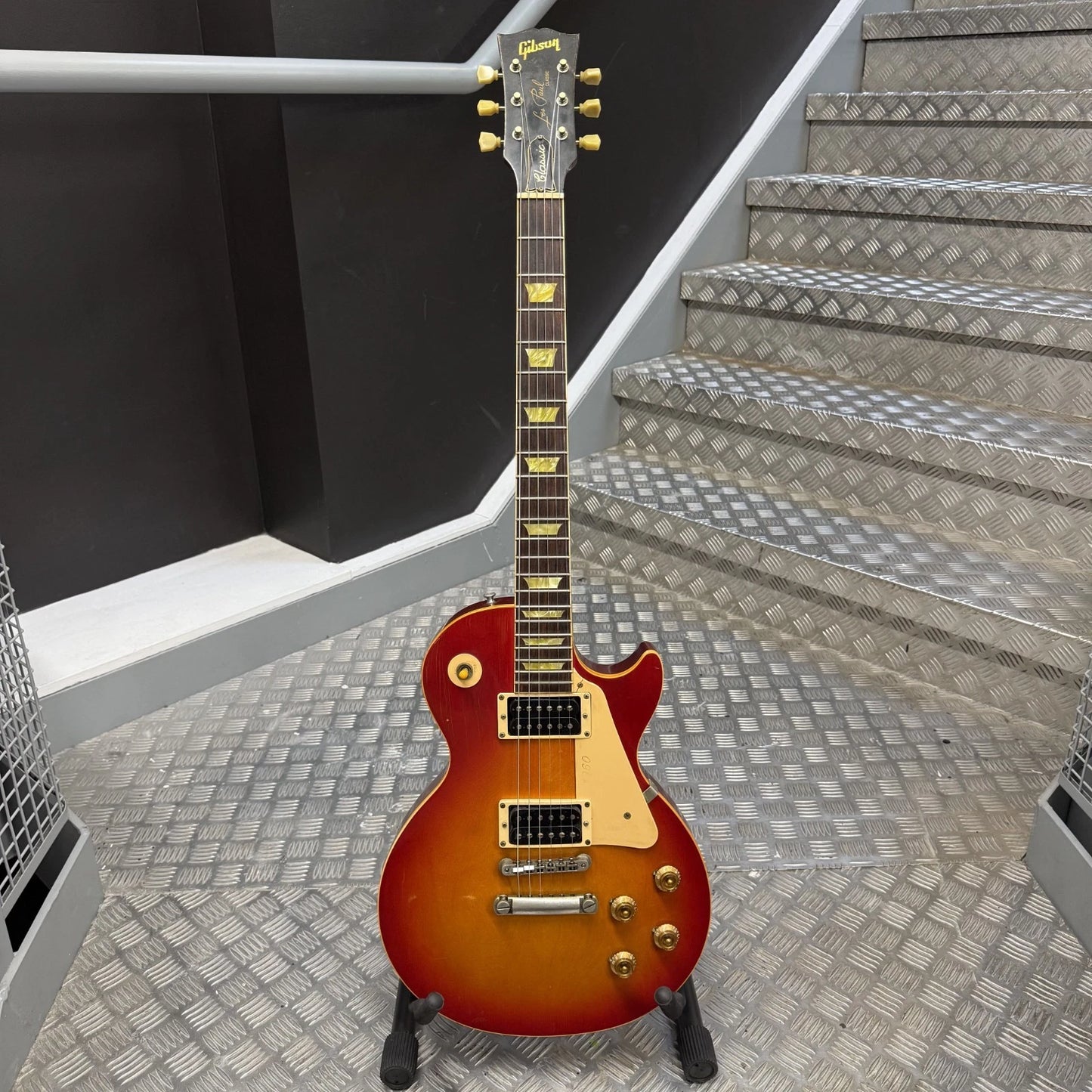 Pre-Owned Gibson Les Paul Classic '1960' 2001 Electric - Cherry Sunburst A Gibson electric guitar with a sunburst finish stands on a stand in front of metal diamond-plate stairs.