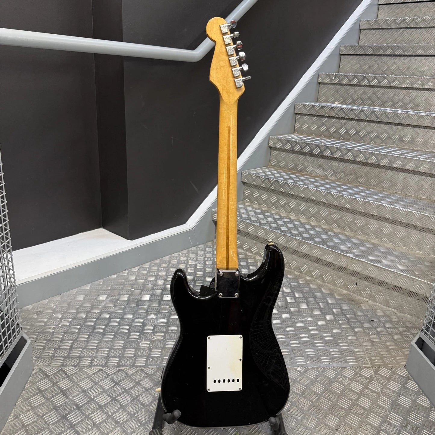Pre-Owned Fender USA American Traditional Fat Stratocaster (Export Model) 1999 Electric - Black A black electric guitar with a natural wood neck, standing on a stand on a diamond-plate metal staircase in an industrial setting.