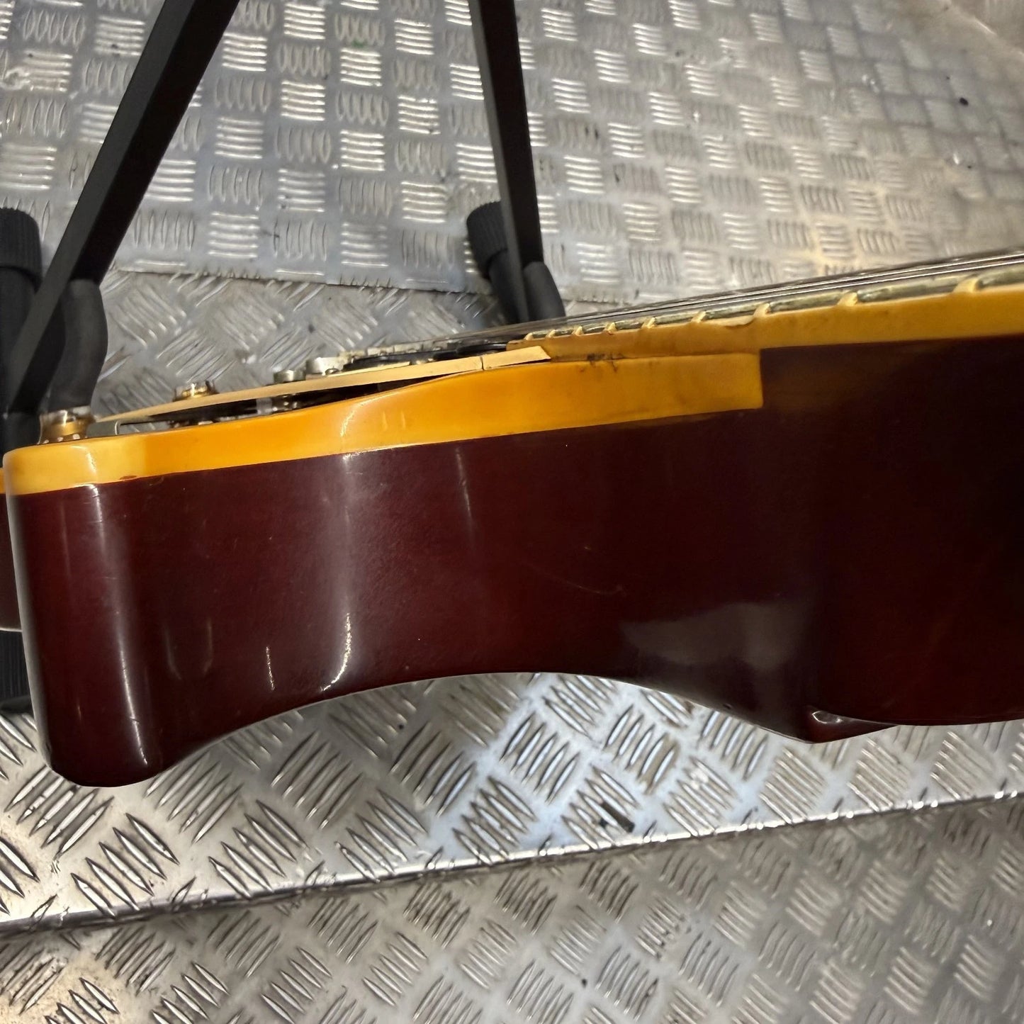 Pre-Owned Gibson Les Paul Classic '1960' 2001 Electric - Cherry Sunburst Close-up of the side of an electric guitar body in glossy dark reddish-brown with a bright yellow binding, resting on a diamond plate metal surface.
