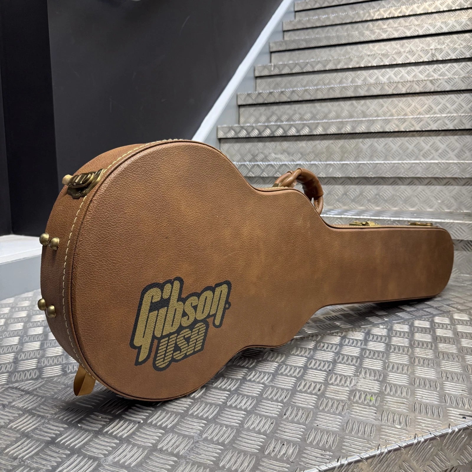 Pre-Owned Gibson Les Paul Classic '1960' 2001 Electric - Cherry Sunburst A brown Gibson guitar case with a textured finish and the 'Gibson USA' logo on the side, resting on a diamond plate metal surface with metal stairs in the background.
