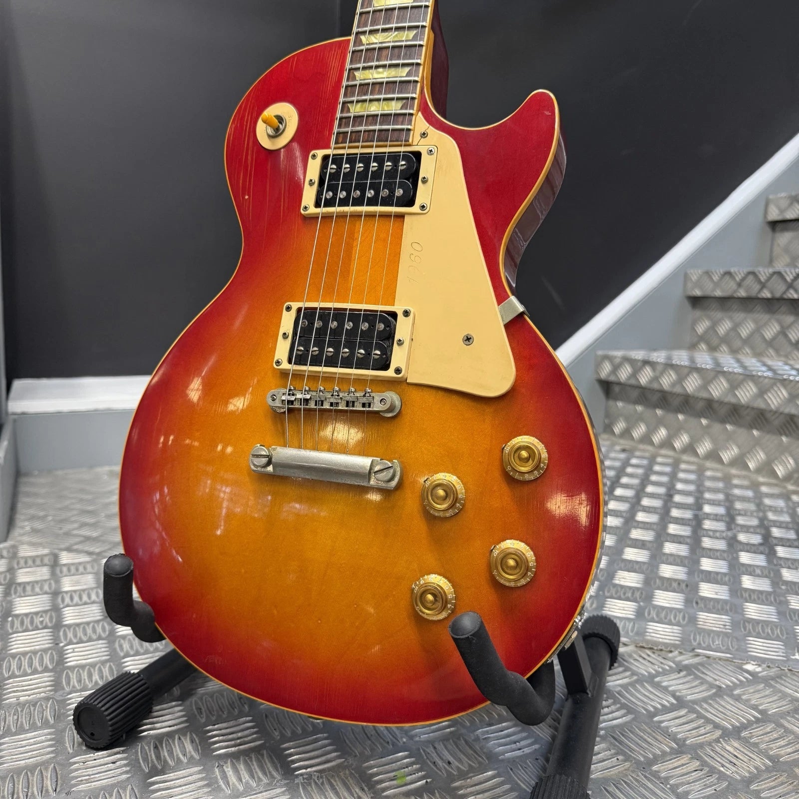 Pre-Owned Gibson Les Paul Classic '1960' 2001 Electric - Cherry Sunburst A Gibson Les Paul Custom electric guitar with a sunburst finish, displayed on a black stand on a diamond plate floor in front of gray steps.